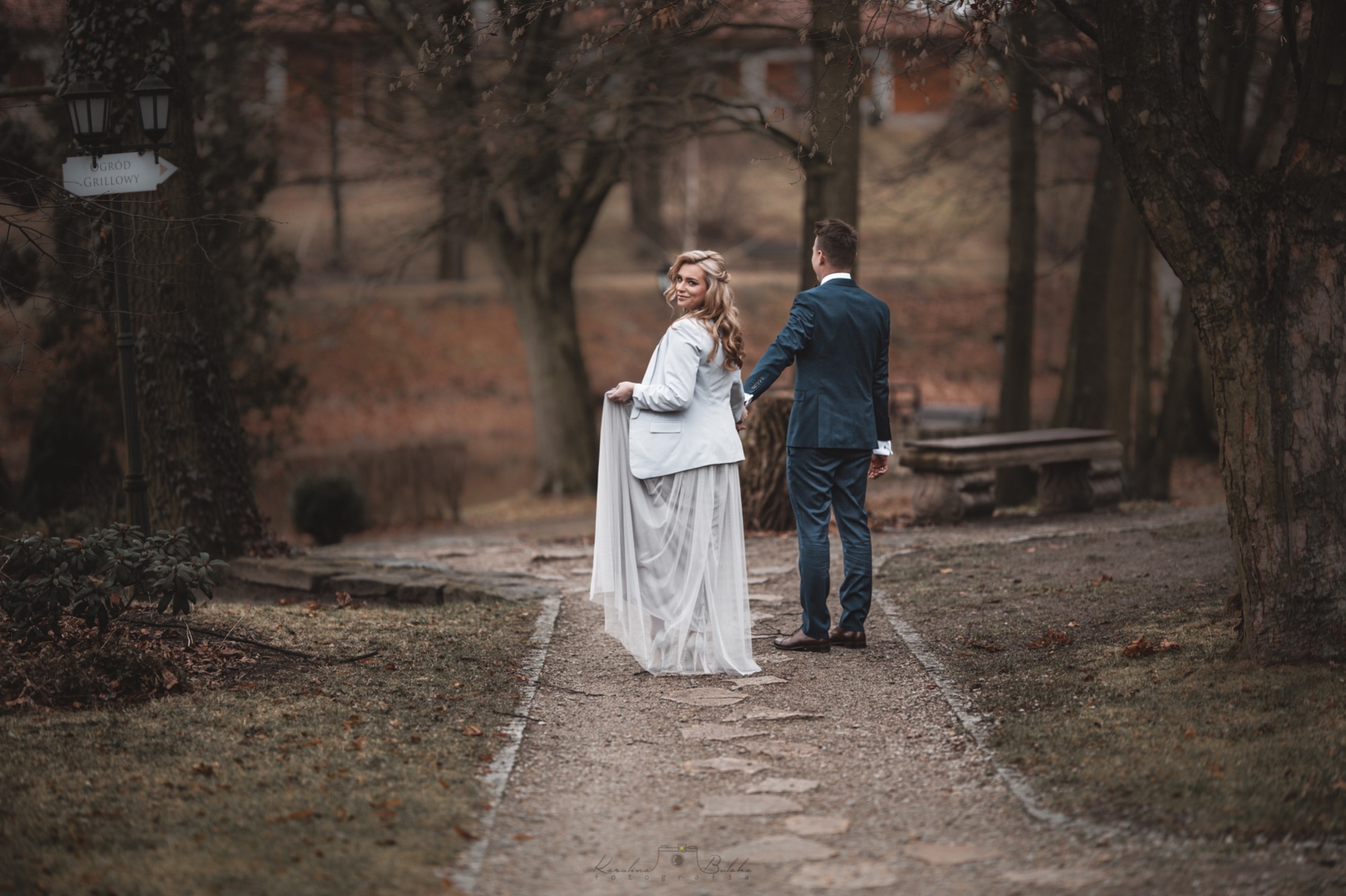 Para w strojach ślubnych spaceruje alejką w parku; panna młoda odwraca się z uśmiechem, trzymając brzeg długiej, zwiewnej spódnicy, a pan młody trzyma ją za rękę; w tle drzewa i drogowskaz.