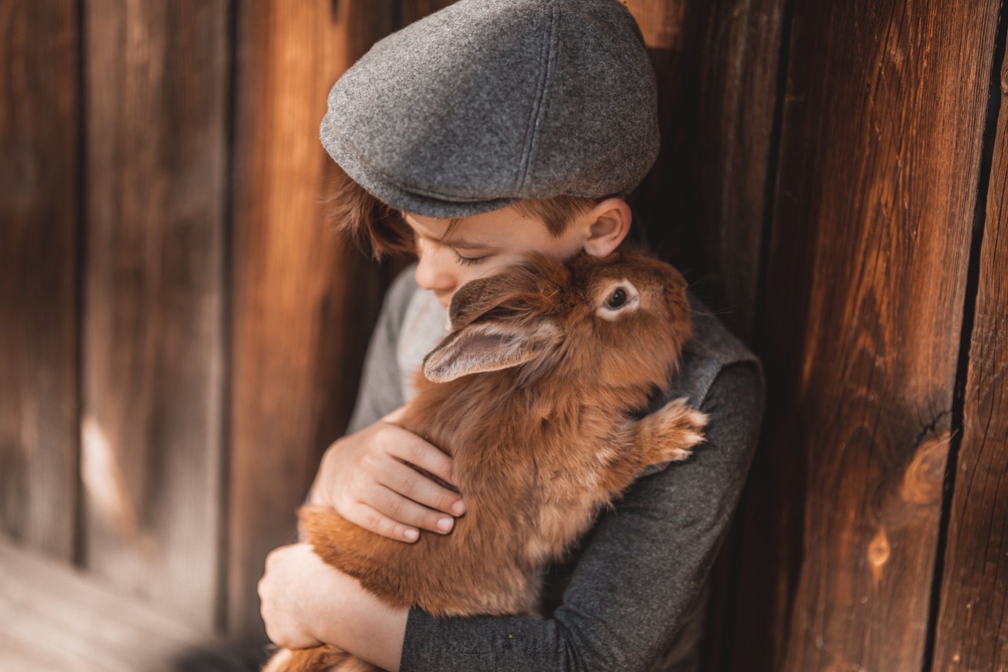 Chłopiec w kaszkiecie przytula brązowego królika na tle drewnianej ściany.