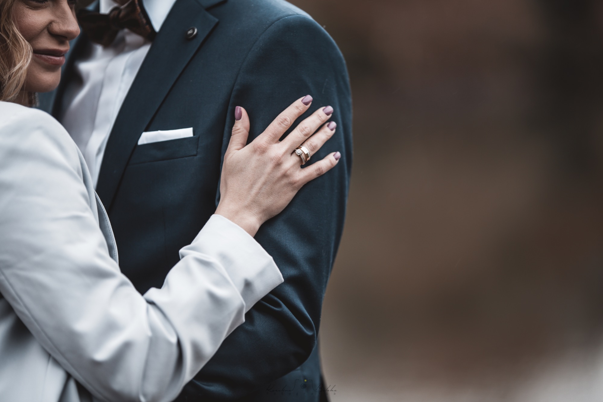 Zbliżenie na parę obejmującą się, widoczne dłonie z obrączkami na tle ciemnego garnituru i jasnej marynarki.
