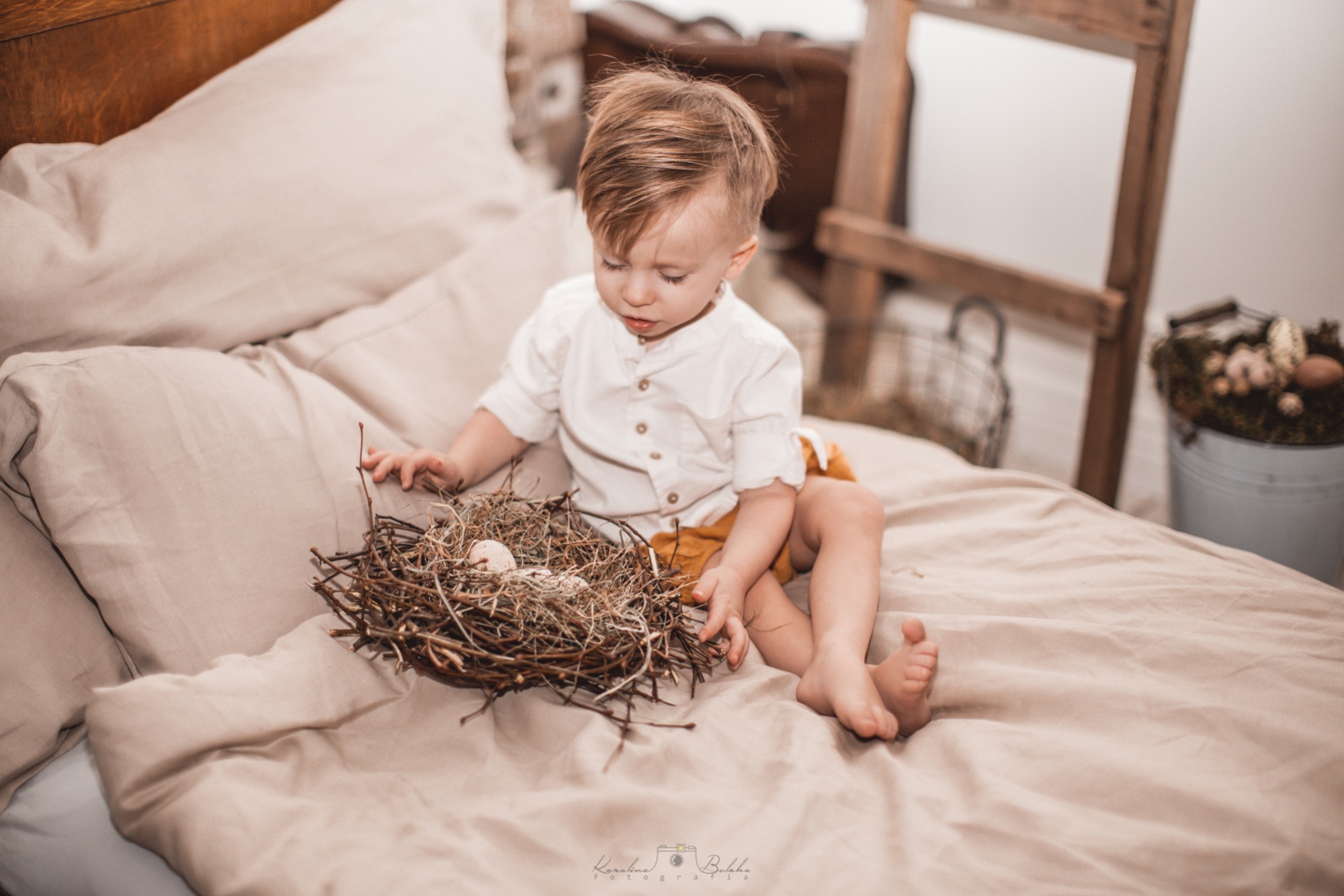 Mały chłopiec w białej koszuli i żółtych spodenkach siedzi na łóżku obok gniazda z dwoma jajkami, z drewnianą drabiną i dekoracyjnym wiaderkiem w tle.