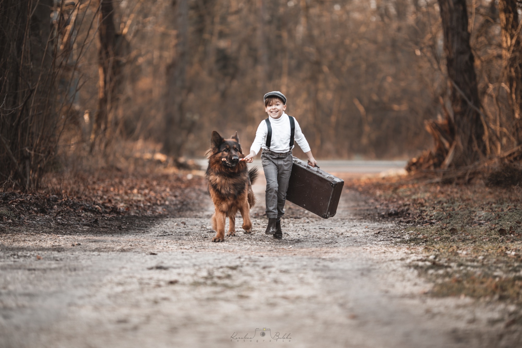 Uśmiechnięty chłopiec w kaszkiecie i szelkach idzie polną drogą, trzymając starą walizkę i prowadząc owczarka niemieckiego.