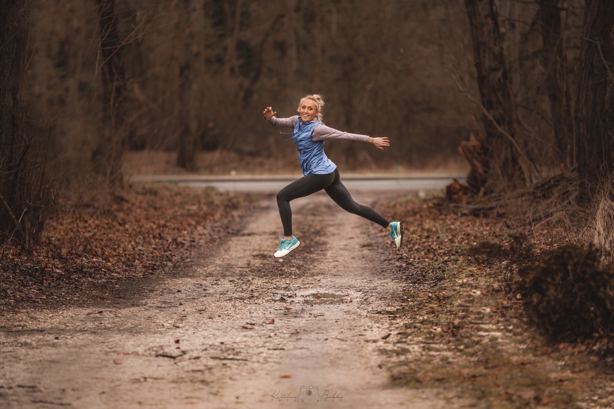 Uśmiechnięta blondynka w sportowym stroju, unosząca się w powietrzu nad leśną ścieżką z kałużami, w dynamicznym skoku.