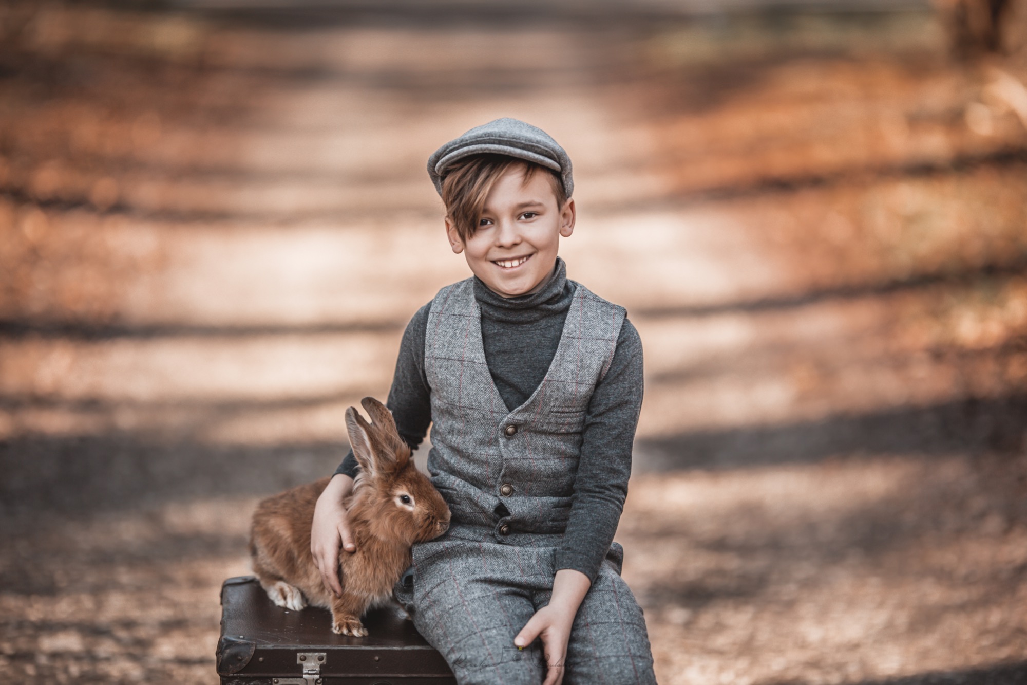 Uśmiechnięty chłopiec w tweedowym garniturze i kaszkiecie siedzi na starej walizce w plenerze, trzymając brązowego królika na kolanach.