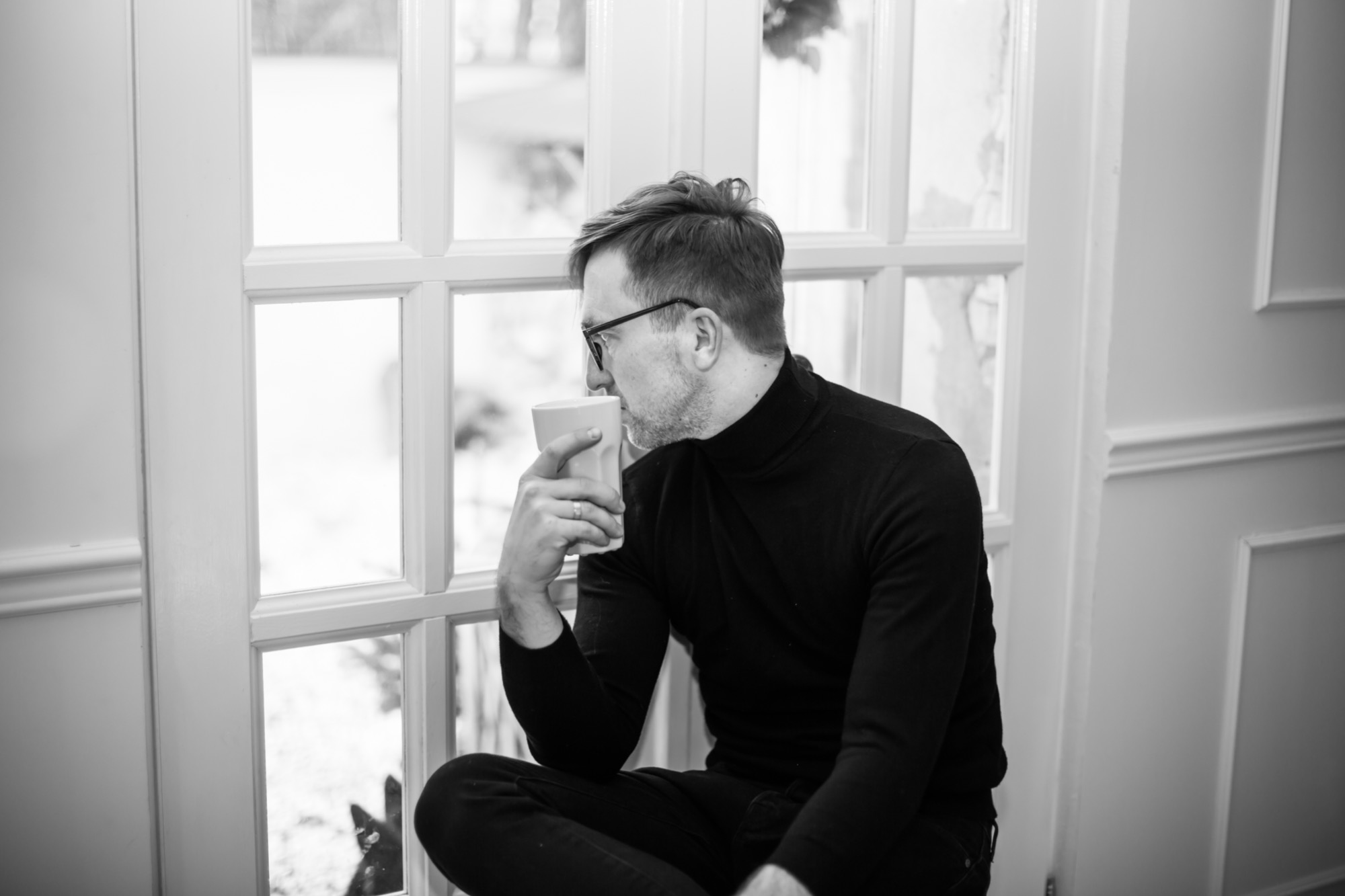 Czarno-białe zdjęcie mężczyzny w okularach, siedzącego na parapecie okna z kubkiem w ręku, patrzącego na zewnątrz.