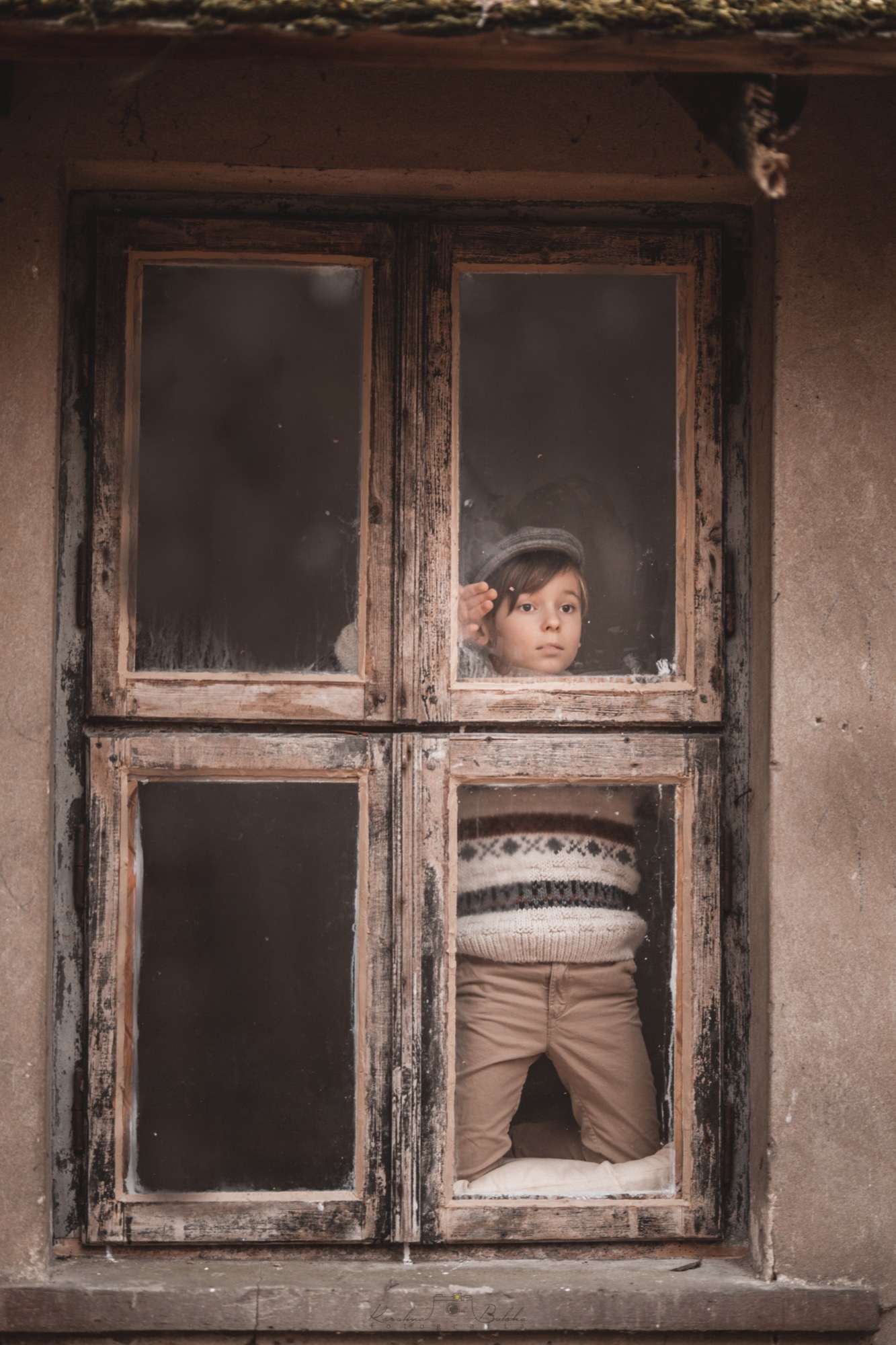 Chłopiec w czapce i swetrze w norweskie wzory patrzy przez stare, drewniane okno z szybami, opierając dłoń o ramę.