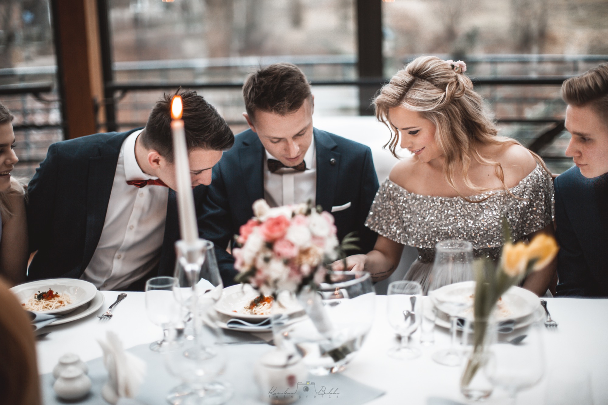 Panna młoda w błyszczącej sukni siedzi przy stole z gośćmi, na stole bukiet kwiatów, świeca i talerze z jedzeniem.