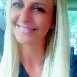 Portret uśmiechniętej kobiety z długimi, platynowymi włosami i niebieskimi oczami, ubrana w ciemny strój, sfotografowana w samochodzie.