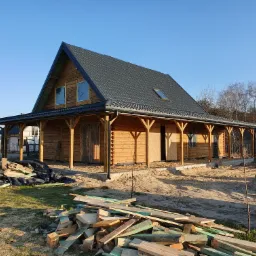 Surowy, drewniany dom z bali z ciemnoszarą dachówką, widoczne materiały budowlane i betoniarka na zewnątrz, w trakcie budowy.