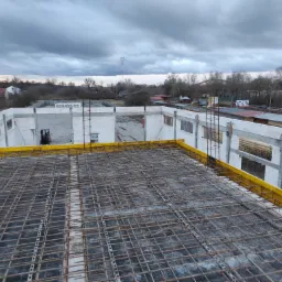 Wiązanie zbrojenia stropu na budowie hali, widoczne szalunki i bloczki Ytong, w tle krajobraz z drzewami i pochmurnym niebem.