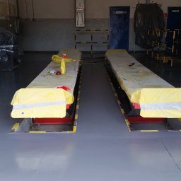 ALTON PIOTR ŁUCZAK - Szara, gładka podłoga w warsztacie samochodowym, z dwoma podnośnikami zabezpieczonymi żółtą folią i taśmą, przygotowanymi do dalszych prac.