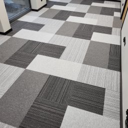 ALTON PIOTR ŁUCZAK - Wnętrze biurowe z ułożoną podłogą z kwadratowych płytek dywanowych w odcieniach szarości i bieli, tworzących geometryczny wzór. Widoczne fragmenty okna i ściany.