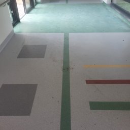 ALTON PIOTR ŁUCZAK - Korytarz w budynku użyteczności publicznej z szarą posadzką w plamy z kolorowymi elementami geometrycznymi (kwadraty, paski), zielonym pasem wzdłuż i zieloną strefą przy oknach i drzwiach.