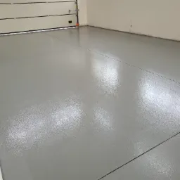 Gładka, szara posadzka w garażu z widocznymi liniami dylatacyjnymi, odbijająca światło, z białą bramą garażową w tle.