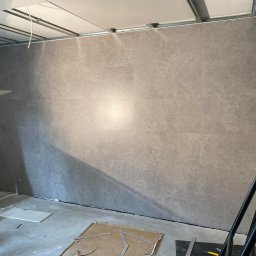 ALTON PIOTR ŁUCZAK - Ściana wyłożona szarymi płytami imitującymi beton, widoczne łączenia, sufit w trakcie montażu, rozstawione rusztowanie i narzędzia na podłodze.