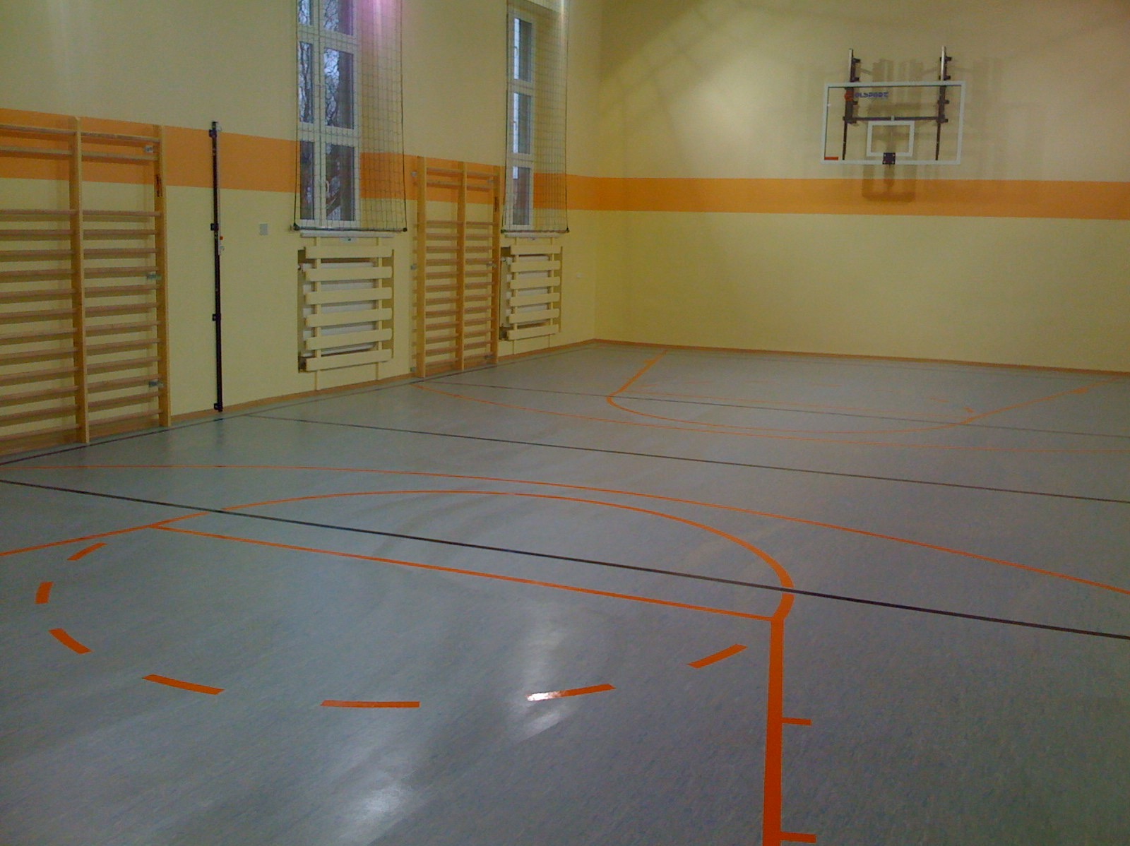 Szara posadzka w hali sportowej z pomarańczowymi liniami boiska, drabinki gimnastyczne przy ścianie i kosz do koszykówki.