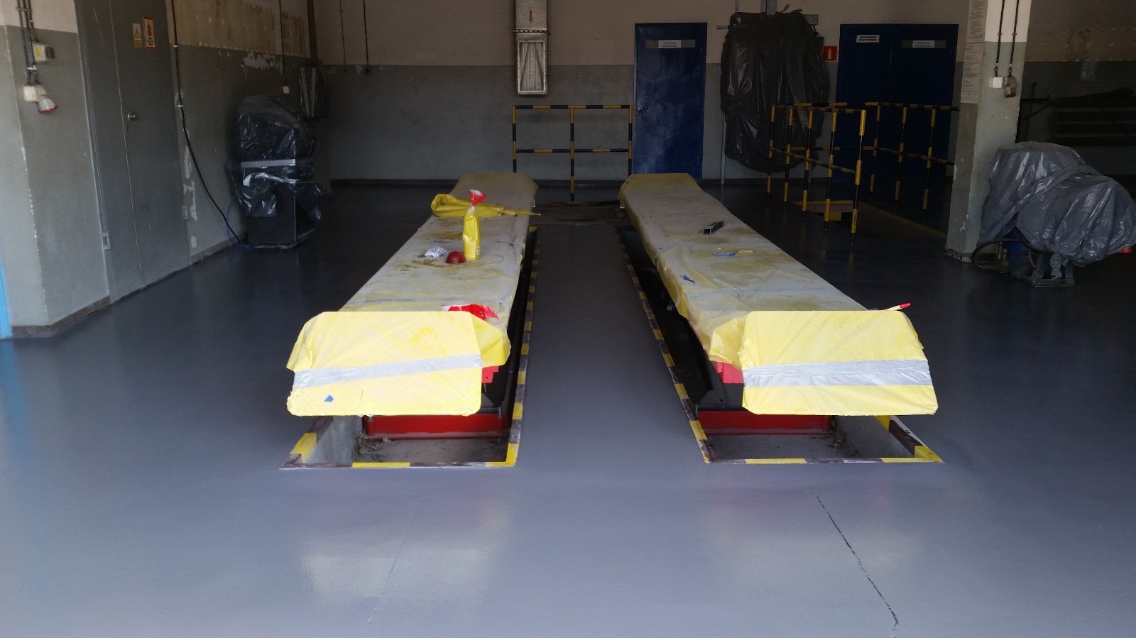 Szara, gładka podłoga w warsztacie samochodowym, z dwoma podnośnikami zabezpieczonymi żółtą folią i taśmą, przygotowanymi do dalszych prac.