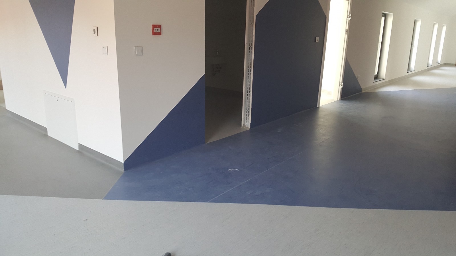 Korytarz w budynku użyteczności publicznej z dwukolorową, szarą i niebieską podłogą, ściany białe z niebieskimi trójkątnymi akcentami, widoczne wejścia do pomieszczeń i okna.