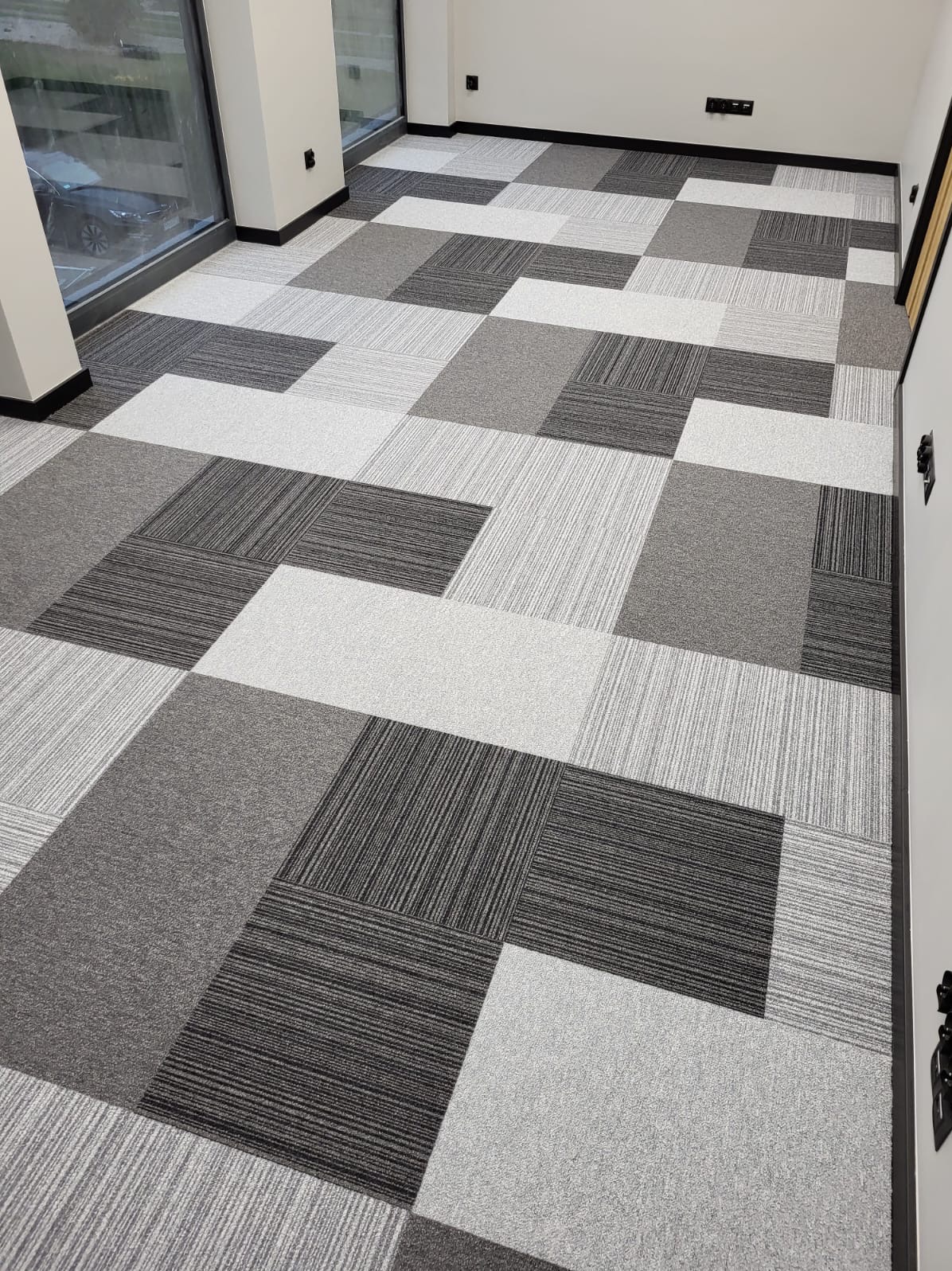 Wnętrze biurowe z ułożoną podłogą z kwadratowych płytek dywanowych w odcieniach szarości i bieli, tworzących geometryczny wzór. Widoczne fragmenty okna i ściany.