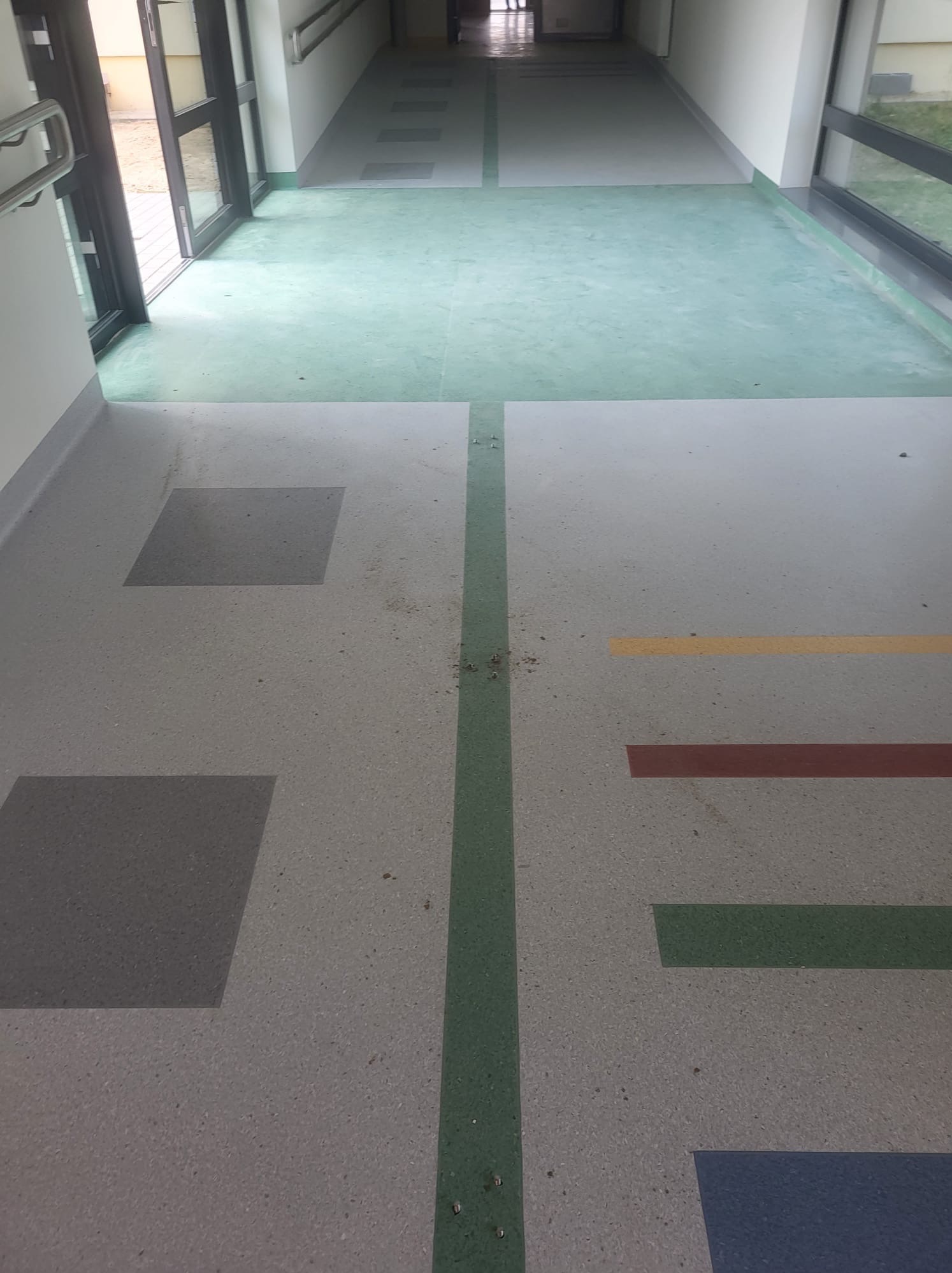 Korytarz w budynku użyteczności publicznej z szarą posadzką w plamy z kolorowymi elementami geometrycznymi (kwadraty, paski), zielonym pasem wzdłuż i zieloną strefą przy oknach i drzwiach.