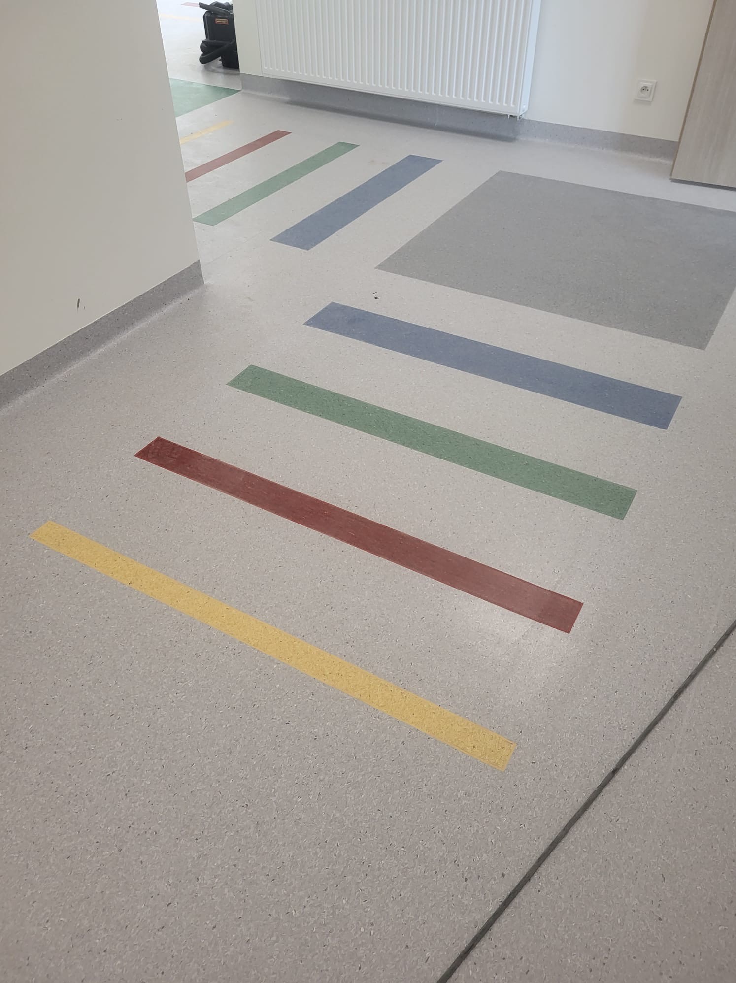 Posadzka żywiczna w korytarzu z kolorowymi pasami (żółty, czerwony, zielony, niebieski) wkomponowanymi w szarą powierzchnię, widoczny grzejnik i fragment ściany.