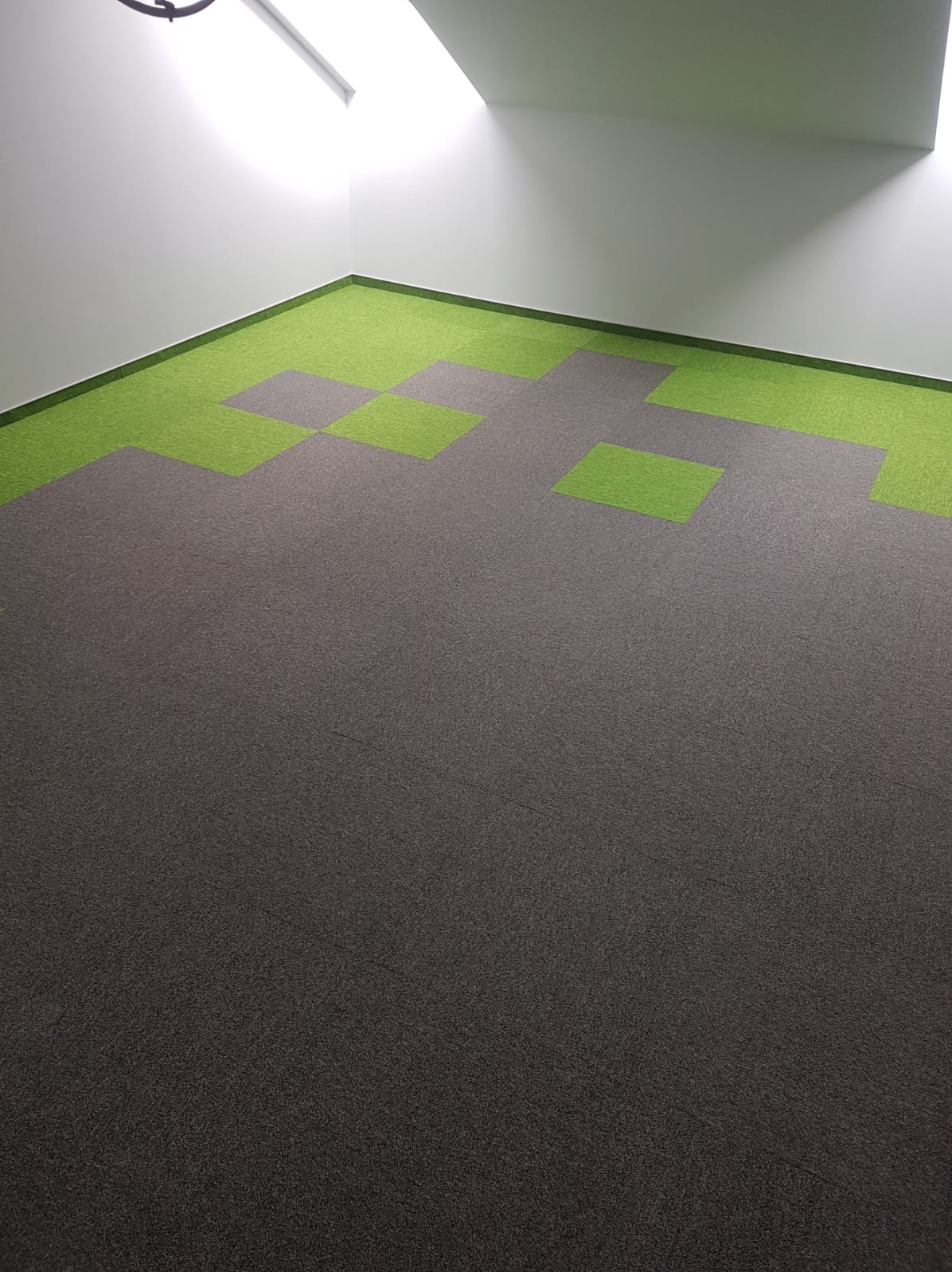 Szara wykładzina dywanowa w biurze z wstawkami z zielonych kwadratów przy ścianach, oświetlona światłem naturalnym wpadającym z góry.