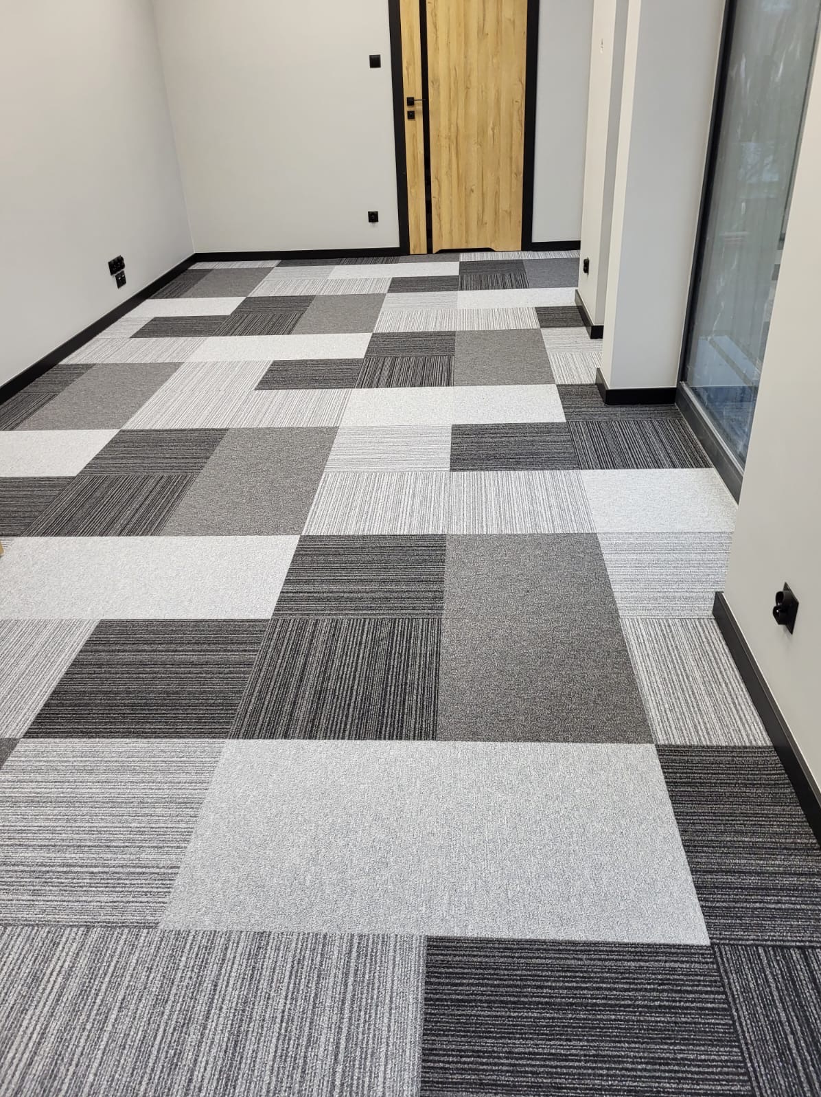 Wnętrze biurowe z podłogą pokrytą kwadratowymi płytkami dywanowymi w odcieniach szarości, ułożonymi w szachownicę; widoczne drzwi z czarną ramą i pionowe okno.