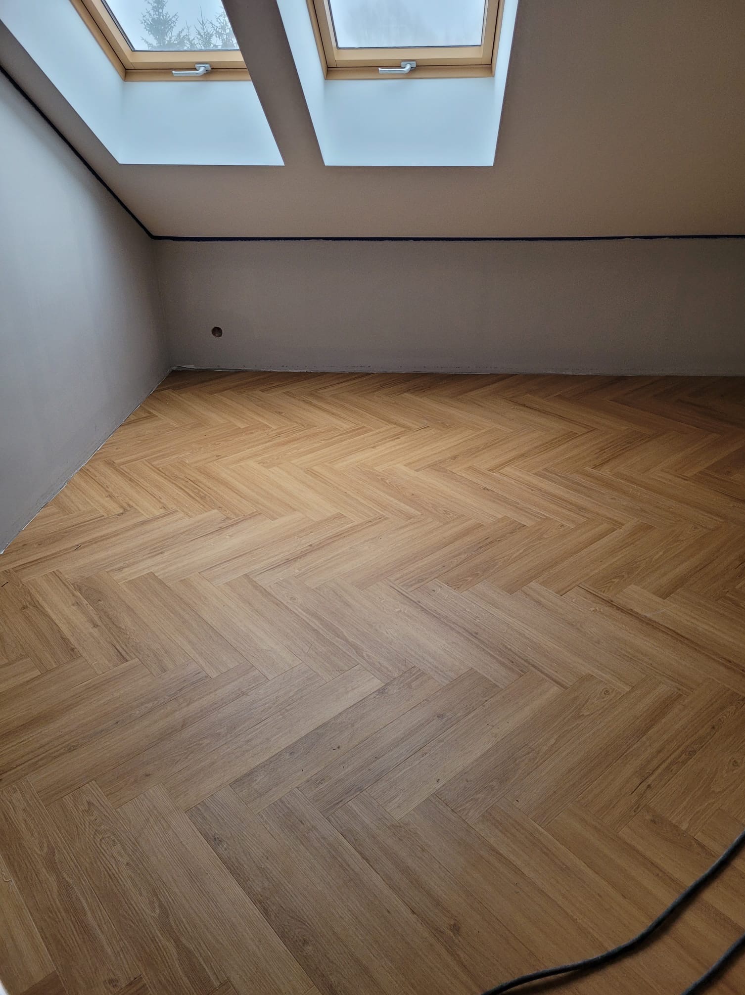 Jasnobrązowa podłoga ułożona w jodełkę w pokoju na poddaszu z dwoma oknami dachowymi, widoczny kabel w prawym dolnym rogu.