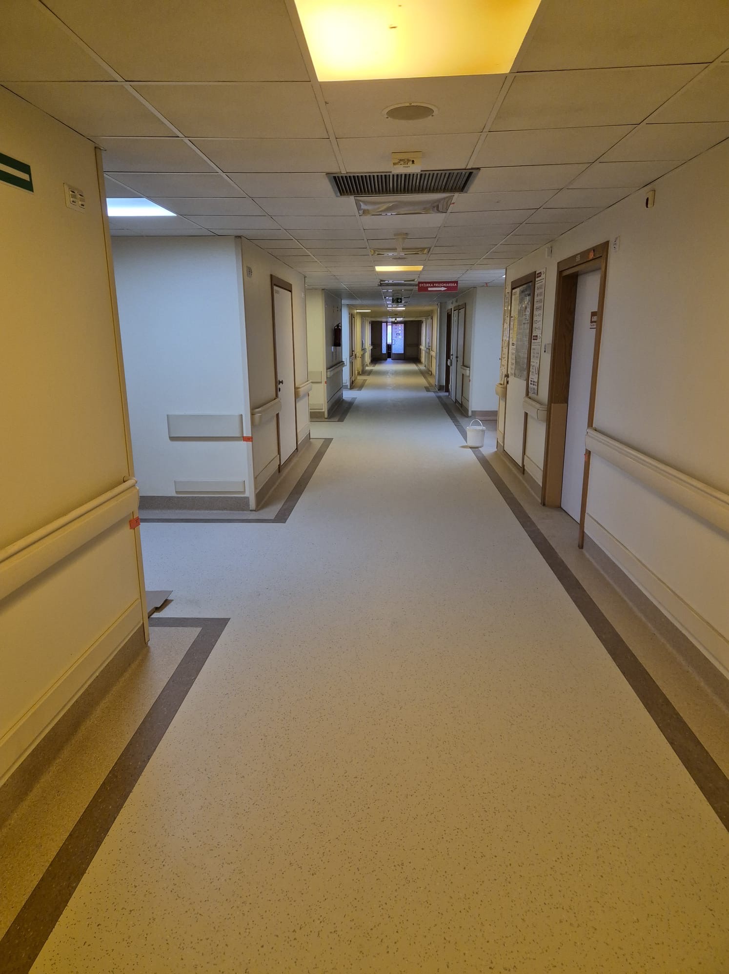 Długi korytarz w budynku użyteczności publicznej z jasną, nakrapianą podłogą i ciemniejszymi pasami przy ścianach, widok w perspektywie.