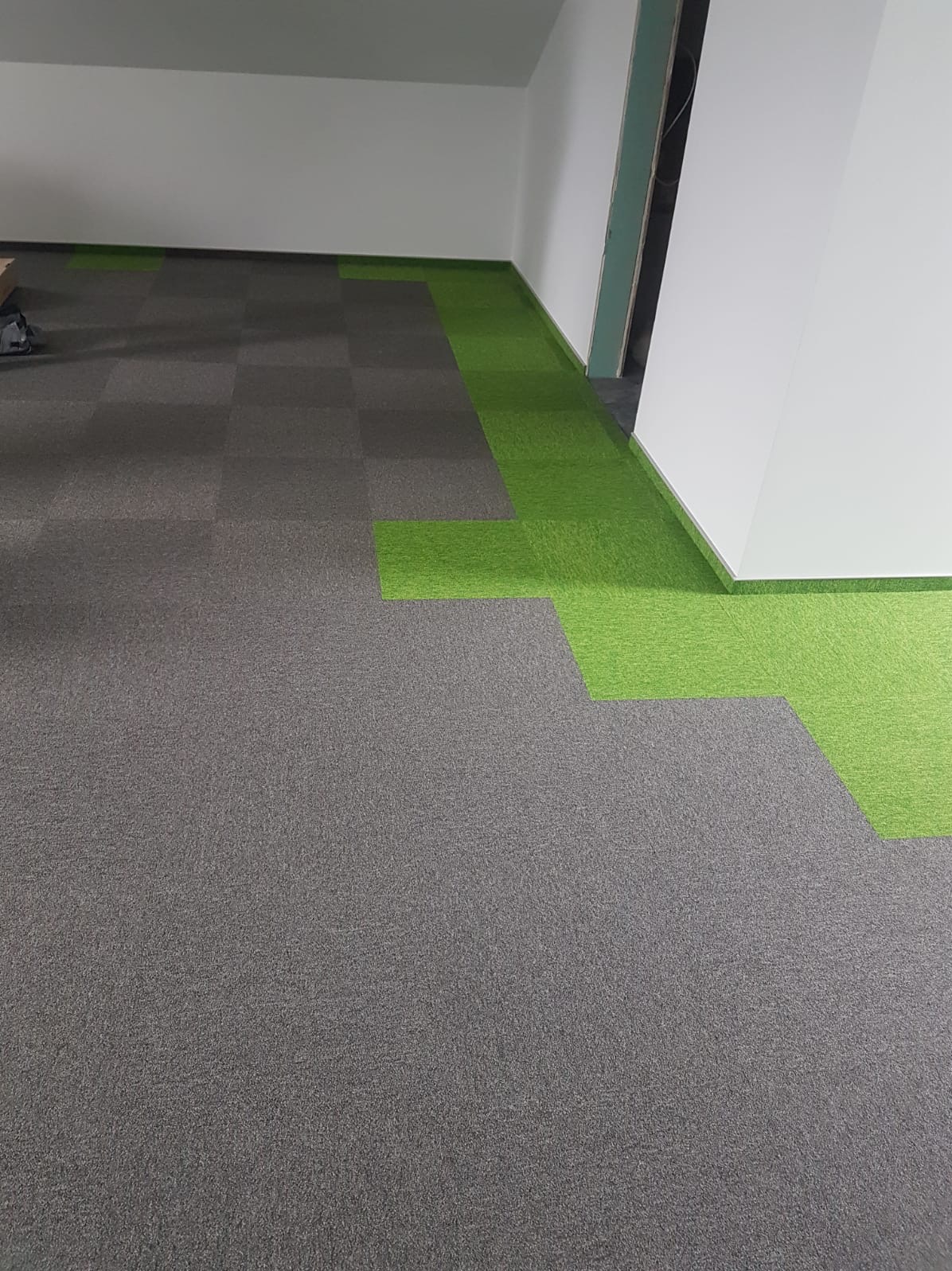 Ułożona w szachownicę szara wykładzina dywanowa w biurze, z fragmentami zielonej wykładziny przy ścianach, tworzącymi obramowanie.
