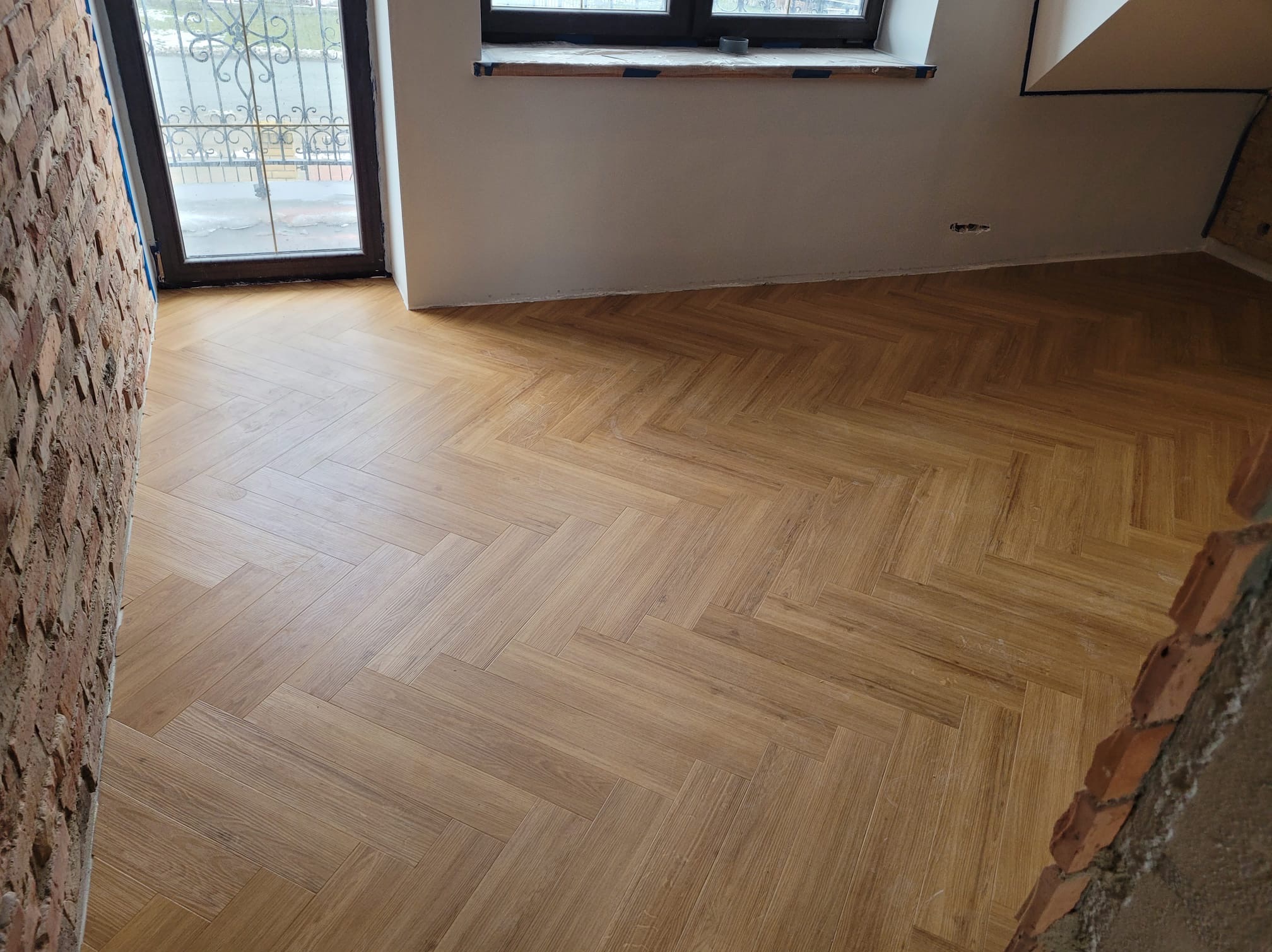 Nowa, jasna podłoga ułożona we wzór jodełki francuskiej w surowym wnętrzu z oknem i fragmentem ceglanej ściany w trakcie wykończenia.