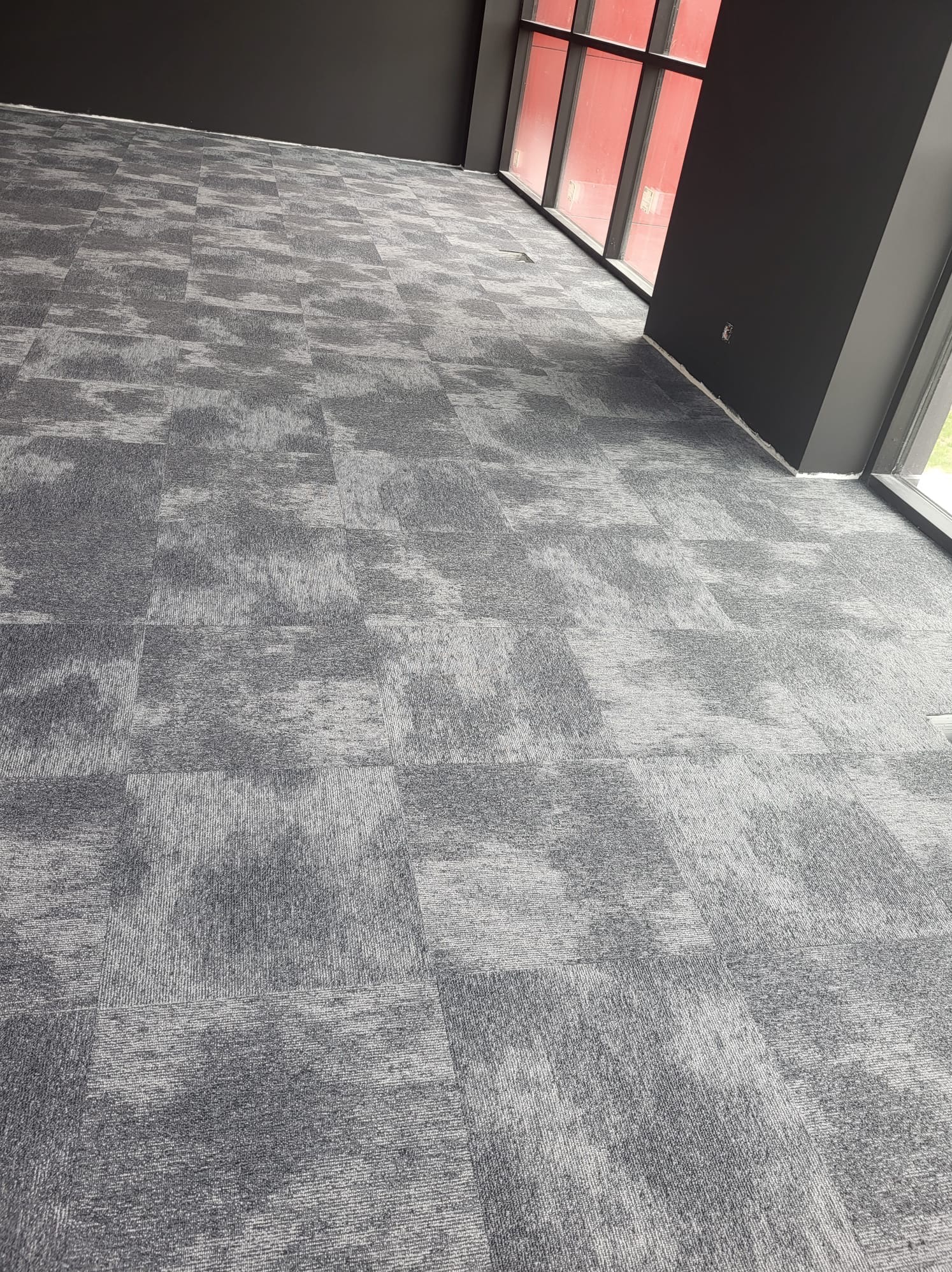 Szara podłoga pokryta kwadratowymi płytkami dywanowymi z delikatnym wzorem, widok z góry w przestronnym wnętrzu z czarnymi ścianami i oknami.