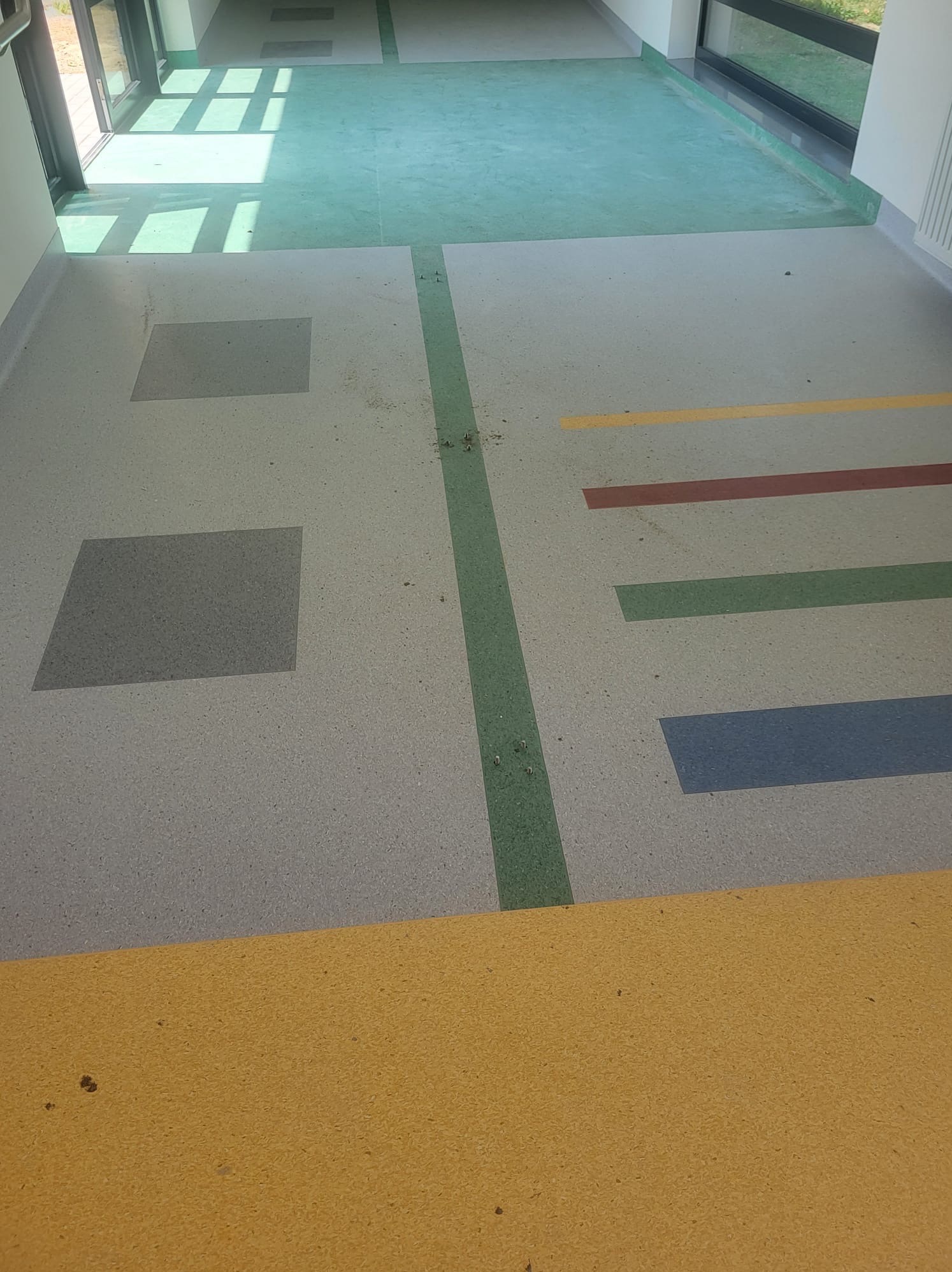 Wykładzina podłogowa w korytarzu z geometrycznymi wzorami w kolorach żółtym, czerwonym, zielonym i niebieskim, z widocznymi śladami po montażu i niedoskonałościami.