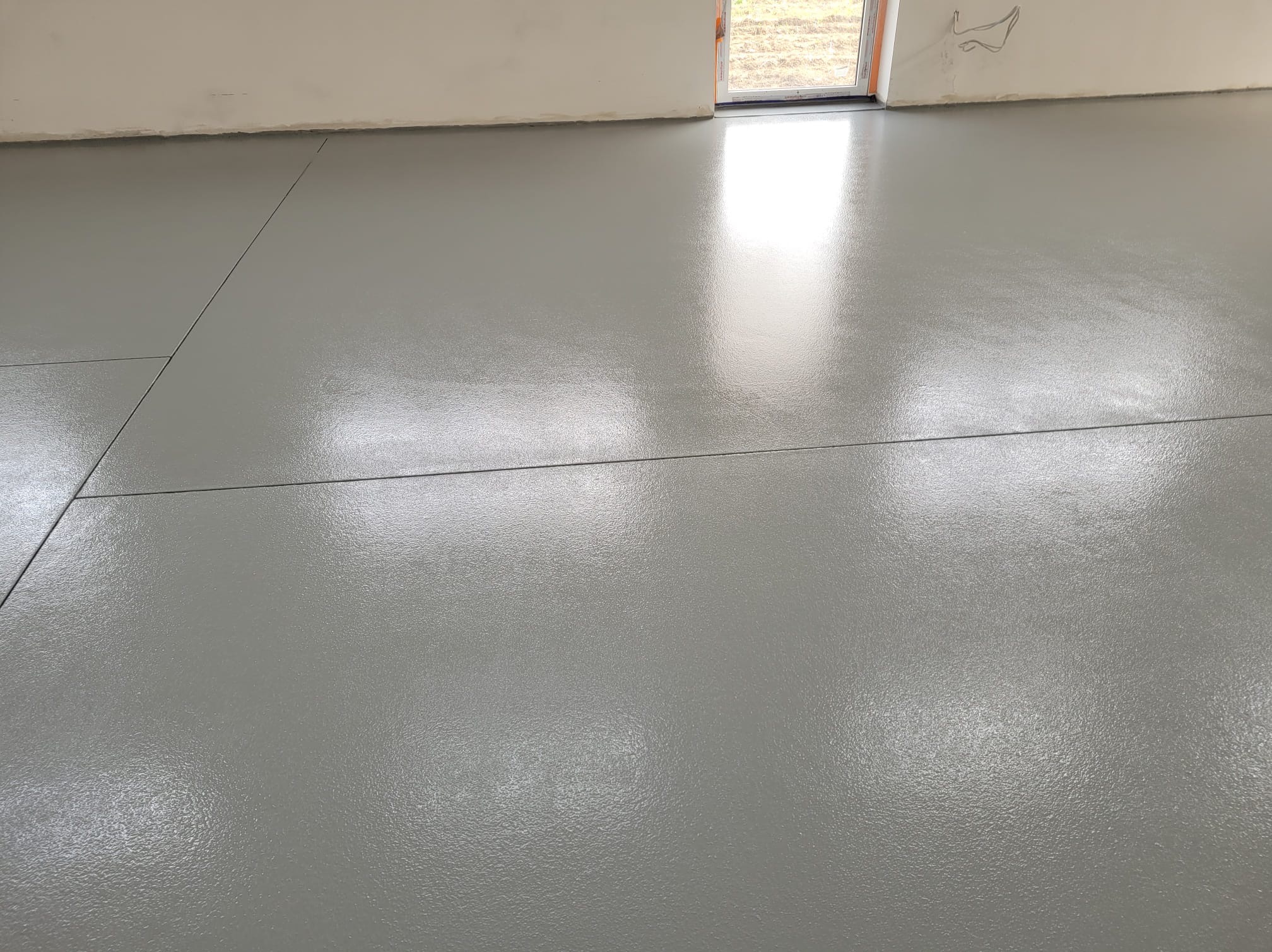 Świeżo wylana, szara posadzka z żywicy w surowym wnętrzu, widoczne dylatacje i odbicie światła z okna.