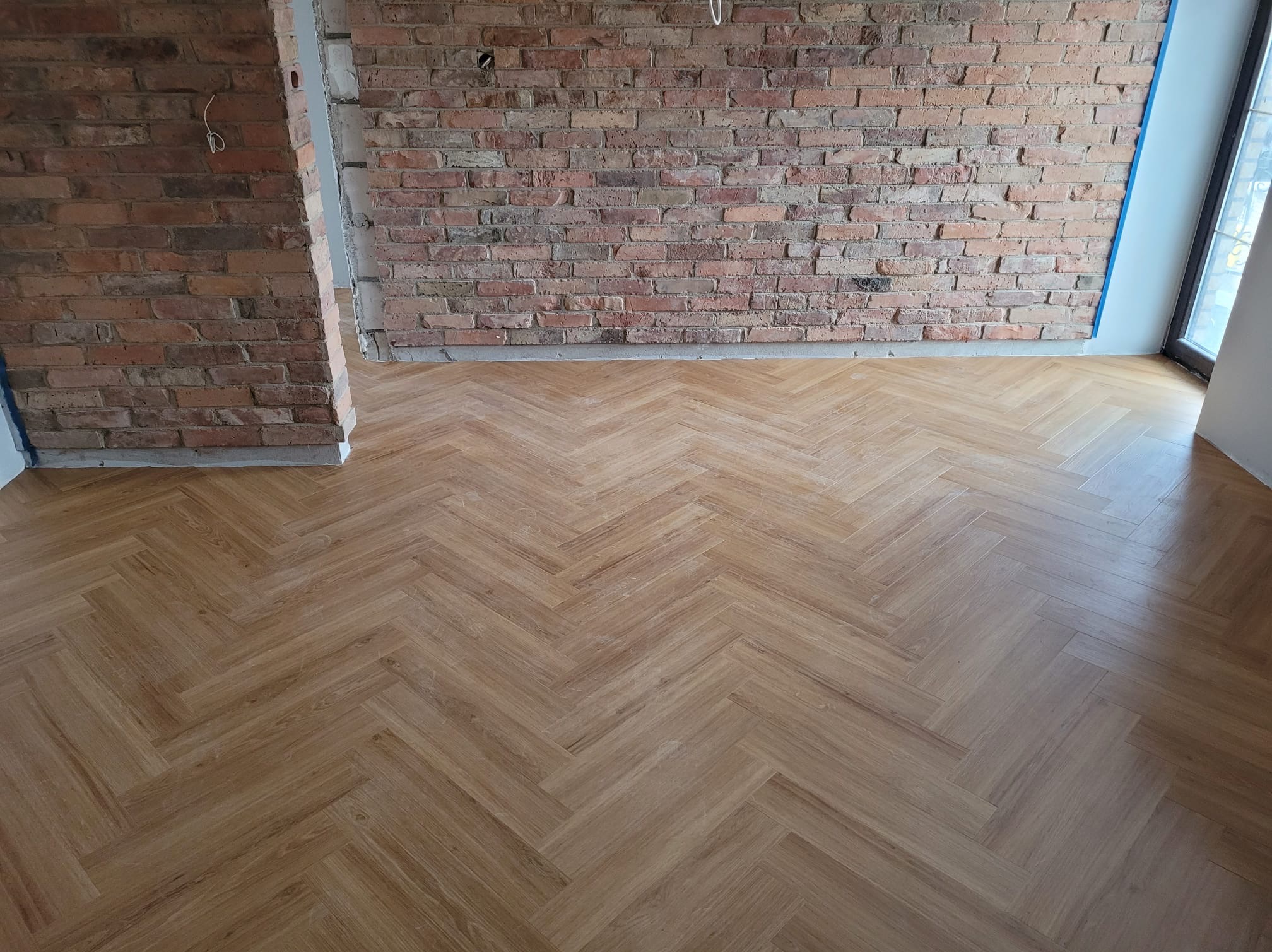 Ułożona podłoga w jodełkę z jasnego drewna, surowa ściana z czerwonej cegły w tle, widoczne ślady po remoncie.