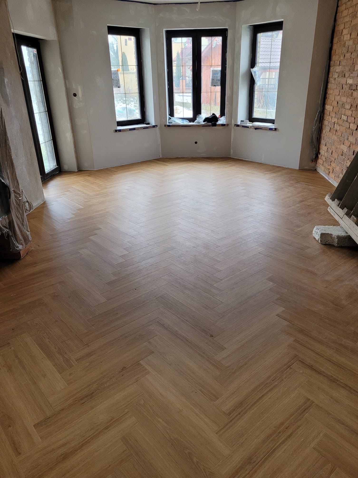 Nowa, jasnobrązowa podłoga ułożona we wzór jodełki w pustym pomieszczeniu z trzema oknami i fragmentem ściany z cegły.