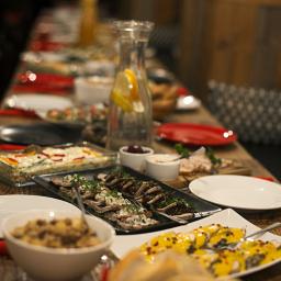 Wilczy Głód - Długi stół z drewnianym blatem zastawiony różnorodnymi daniami: sałatki w szklanym naczyniu, mięsa na ciemnym półmisku, żółte roladki, białe miseczki z sosami, dzbanek z wodą i cytryną, czerwone...