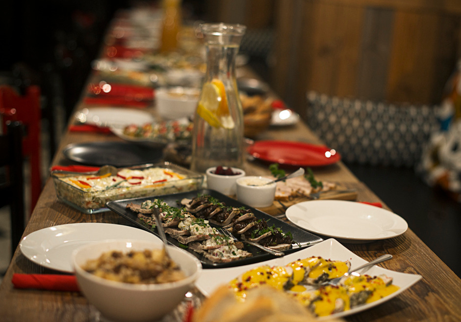 Długi stół z drewnianym blatem zastawiony różnorodnymi daniami: sałatki w szklanym naczyniu, mięsa na ciemnym półmisku, żółte roladki, białe miseczki z sosami, dzbanek z wodą i cytryną, czerwone...