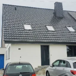 Dom jednorodzinny z ciemnoszarą dachówką, trzema oknami dachowymi i panelami słonecznymi na dachu, widok z zewnątrz na podjazd z zaparkowanymi samochodami i budynkiem garażowym z niebieską bramą.