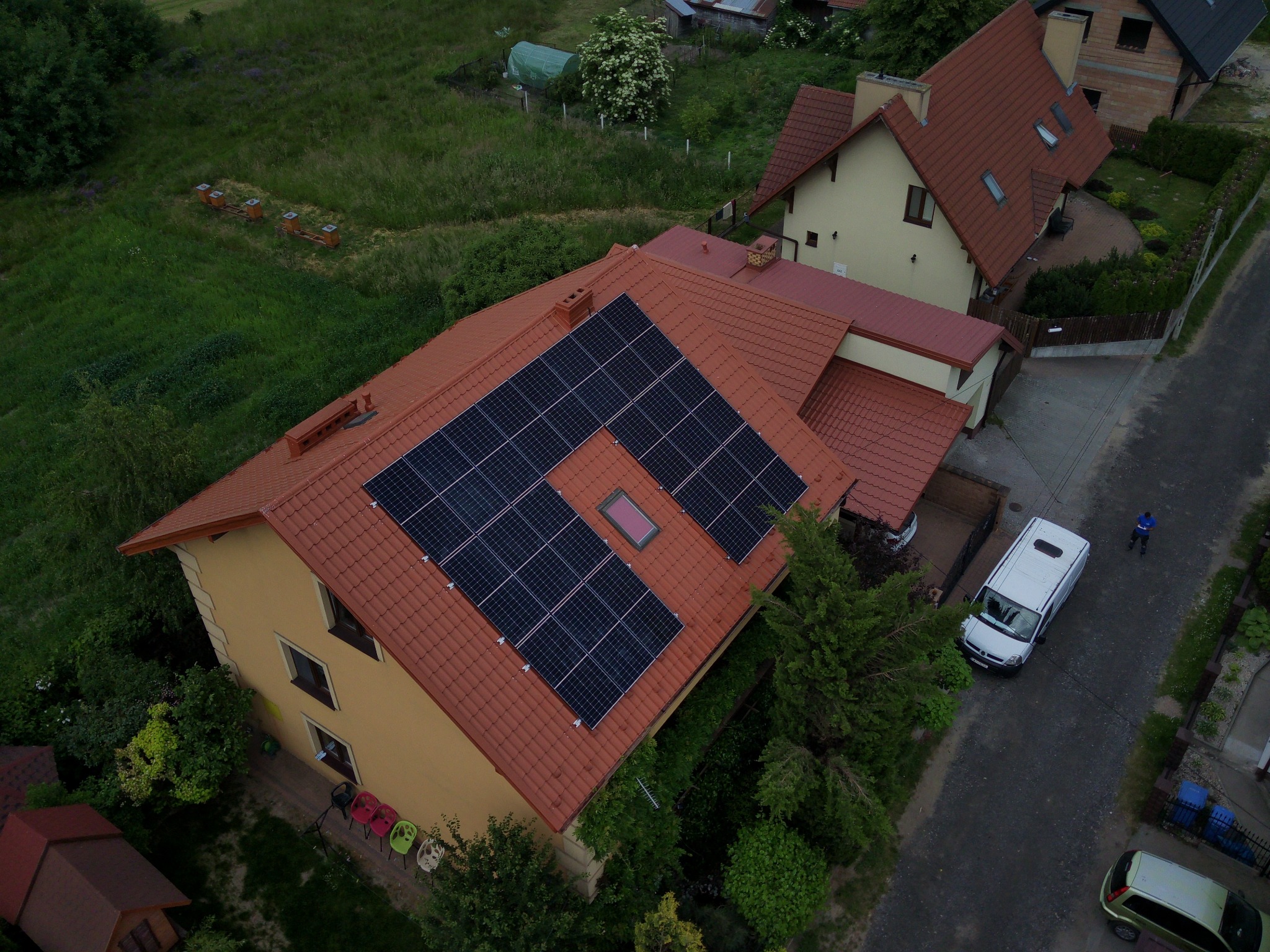 Widok z drona na dom z panelami słonecznymi na czerwonym dachu, obok zaparkowany biały bus, w tle ule dla pszczół.