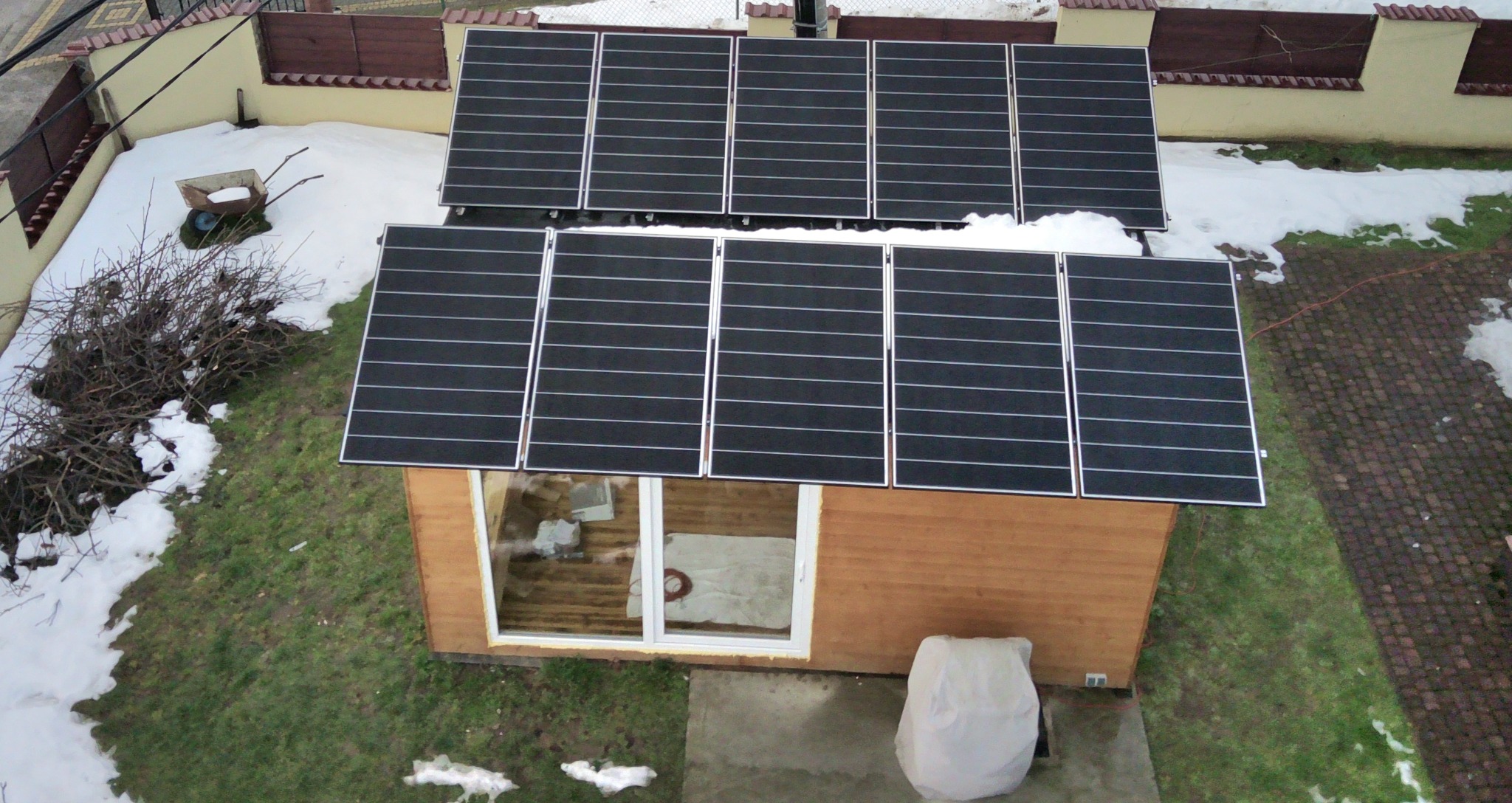 Widok z góry na mały, drewniany budynek z panelami fotowoltaicznymi na dachu, częściowo pokrytym śniegiem, otoczony trawnikiem z resztkami śniegu i drzewami bez liści.