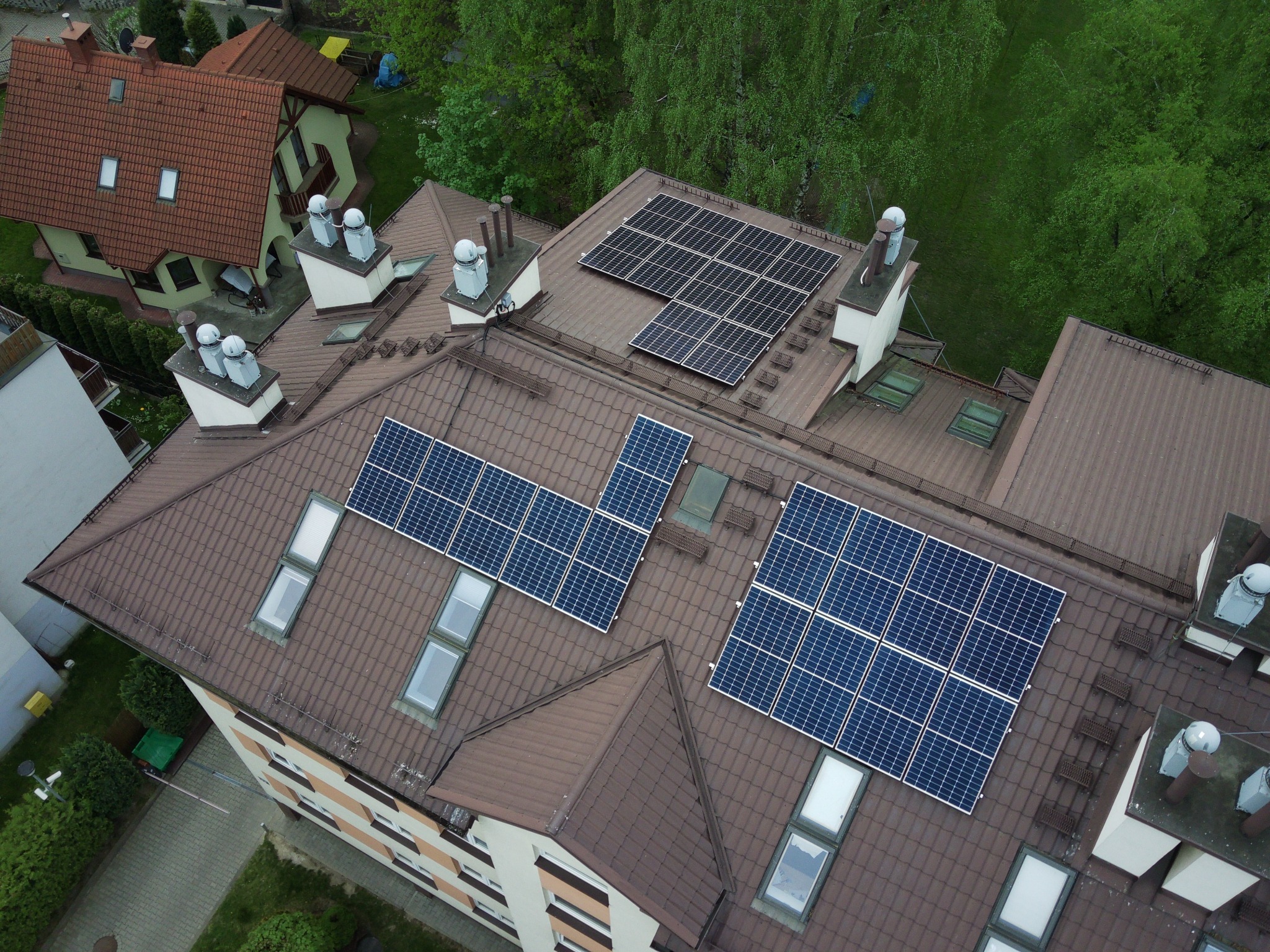 Widok z góry na brązowy dach wielorodzinnego budynku z zainstalowanymi panelami słonecznymi, kominami i oknami dachowymi, otoczony zielenią i innymi budynkami.