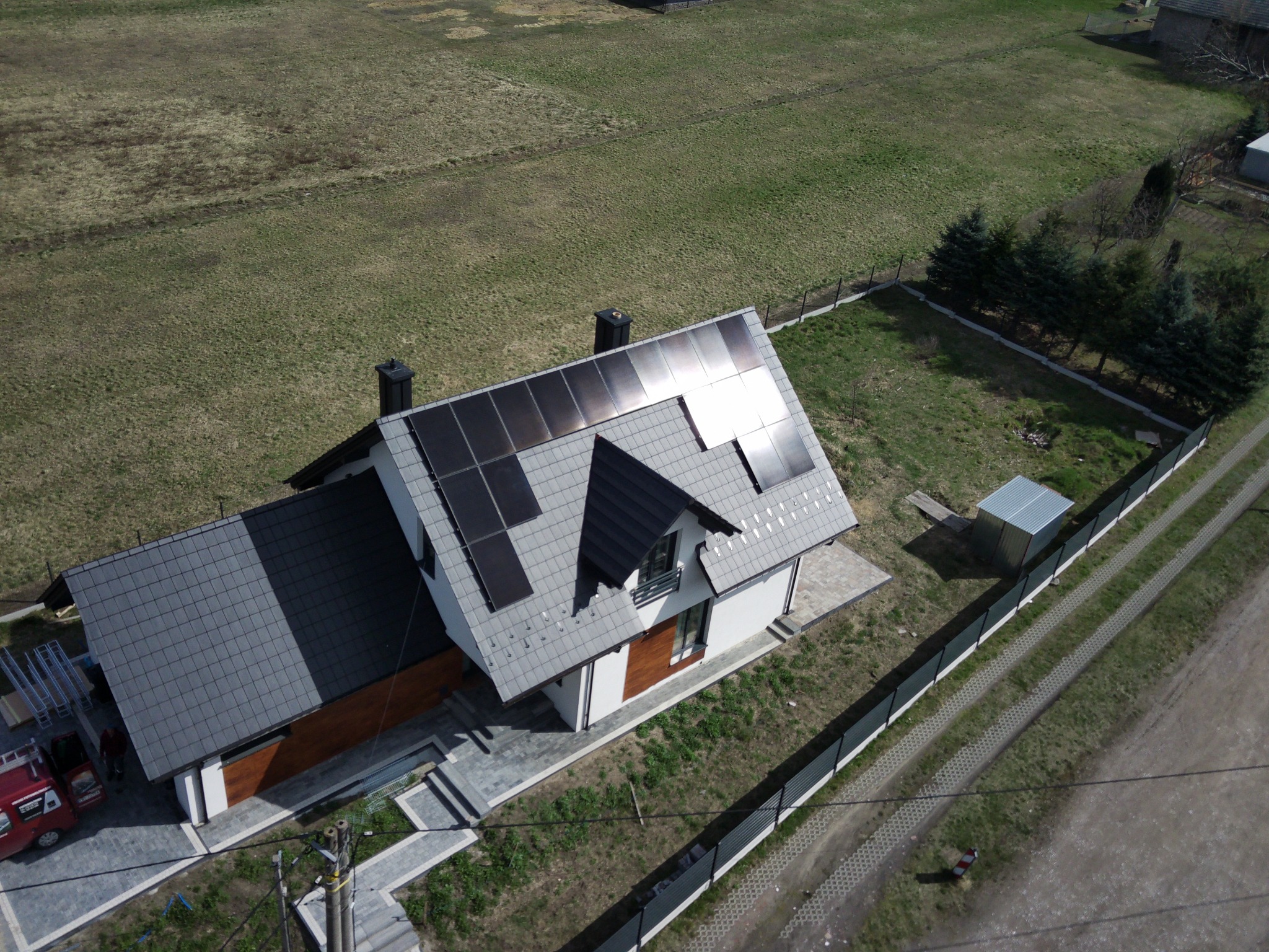 Widok z drona na dom z szarym dachem i panelami słonecznymi, z czerwonym samochodem dostawczym i elementami montażowymi obok, otoczony trawnikiem i ogrodzeniem.