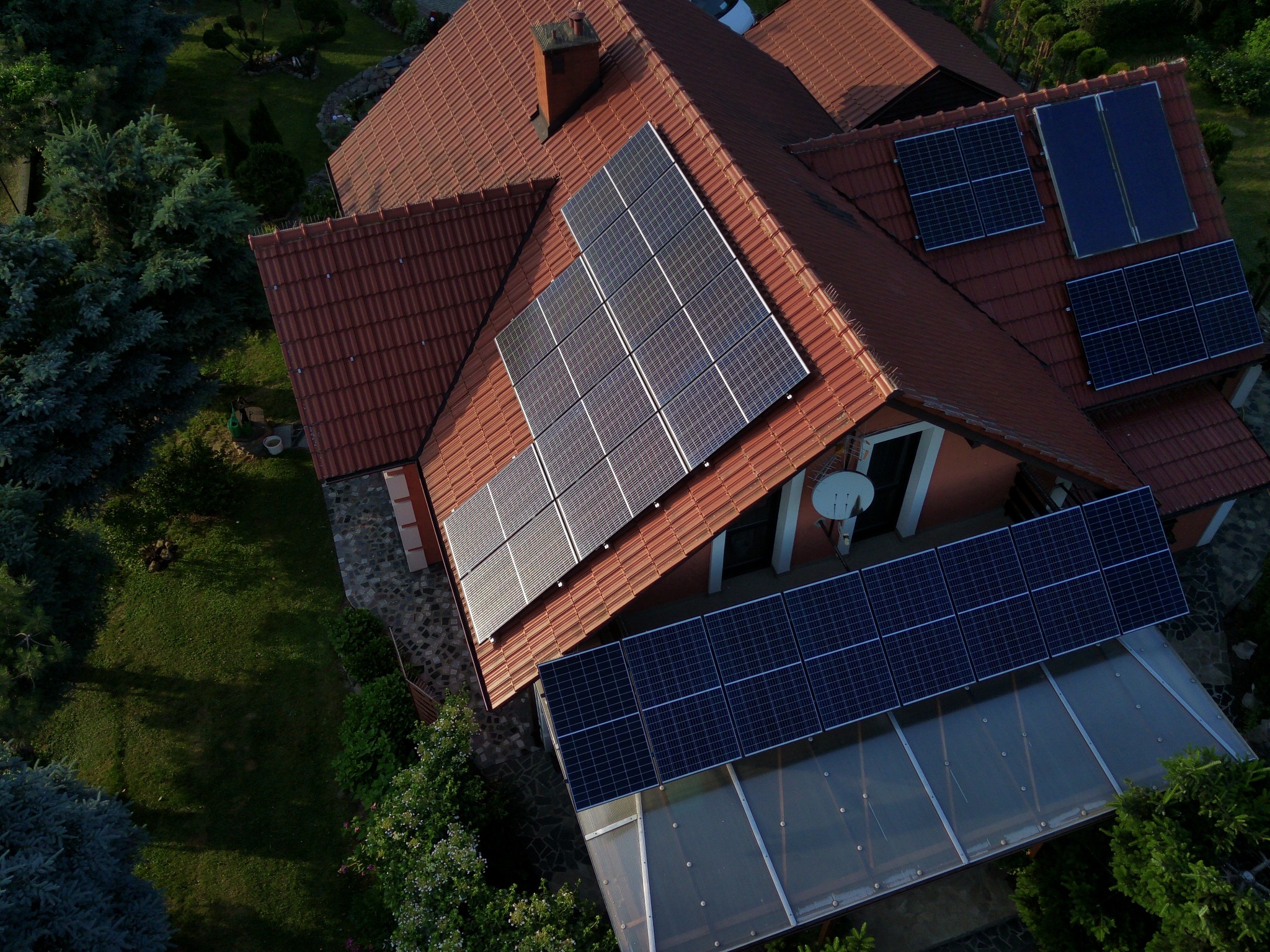 Widok z góry na dom z czerwoną dachówką, na której zamontowane są panele słoneczne; część paneli znajduje się nad przeszkloną werandą