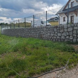 Bartłomiej Fiedor - Granitowa ściana oporowa z siatkowym ogrodzeniem na tle domu i pochmurnego nieba, z widocznym systemem nawadniania trawnika.