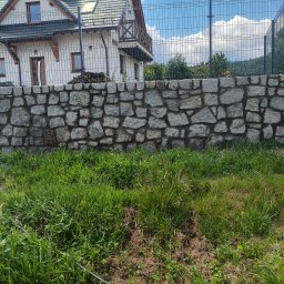 Bartłomiej Fiedor - Murek z nieregularnych kamieni granitowych przed domem z balkonem, za ogrodzeniem z siatki.