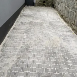 Ułożona szara kostka brukowa typu I na wąskim chodniku między szarą ścianą budynku a kamiennym murem. Widok z góry, lekko z ukosa.