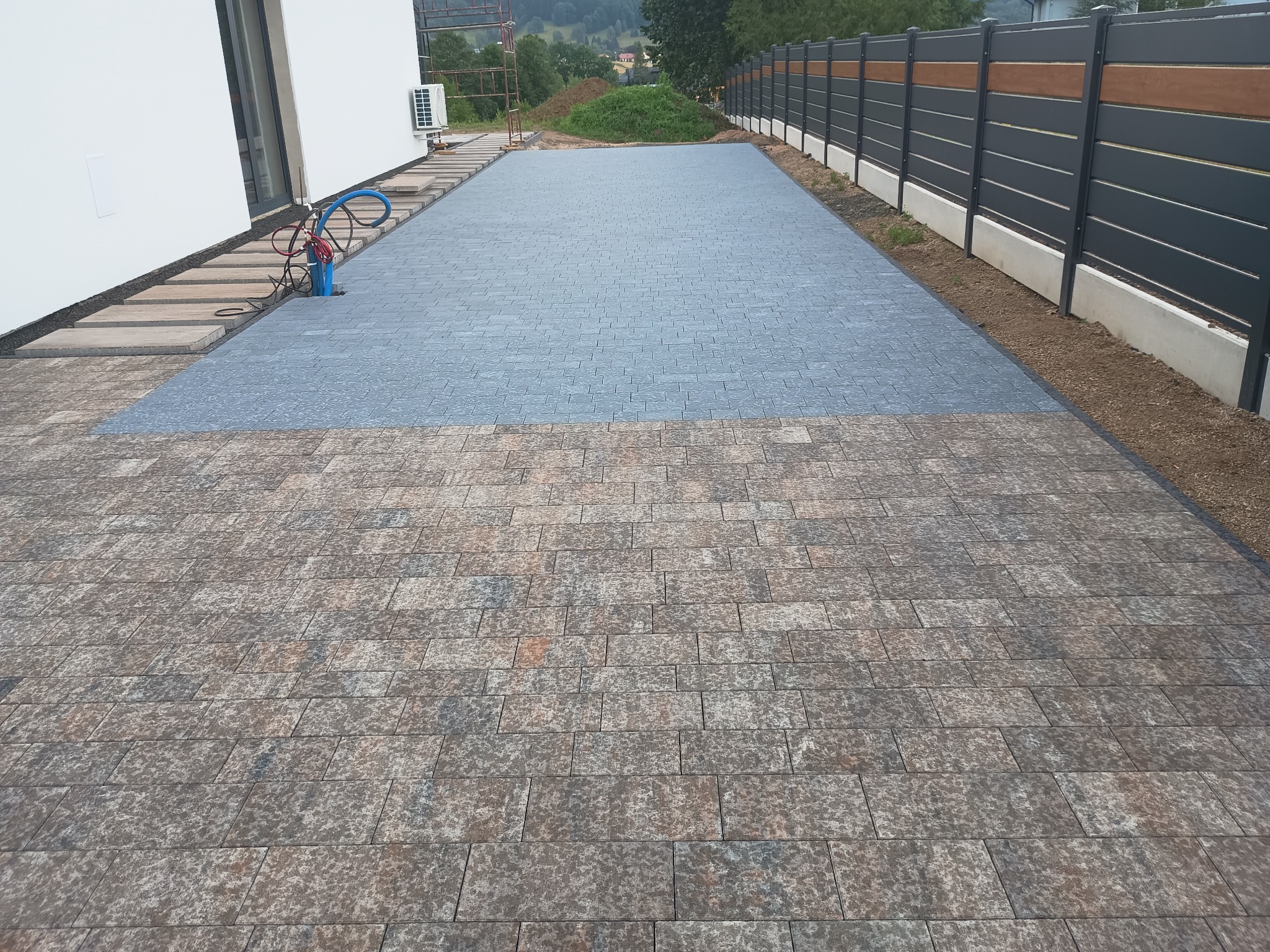 Ułożona kostka brukowa na podjeździe z widocznym fragmentem nowej, szarej nawierzchni oraz starszej, brązowo-szarej kostki, obok domu z klimatyzatorem i nowoczesnym ogrodzeniem panelowym.