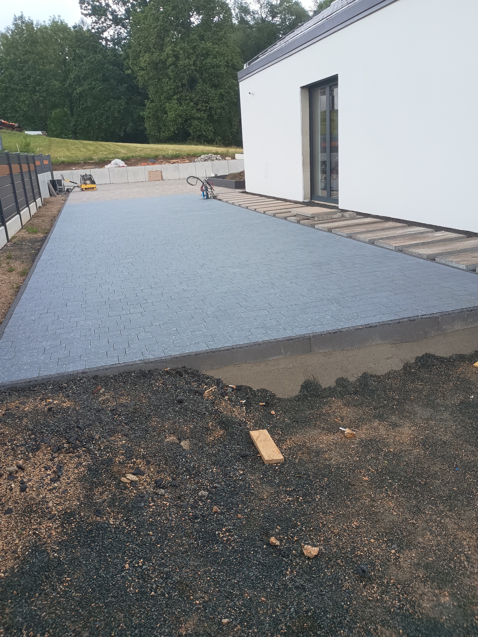 Świeżo ułożona, szara kostka brukowa przed nowoczesnym domem z białymi ścianami, widoczne przygotowane podłoże i narzędzia brukarskie w tle.