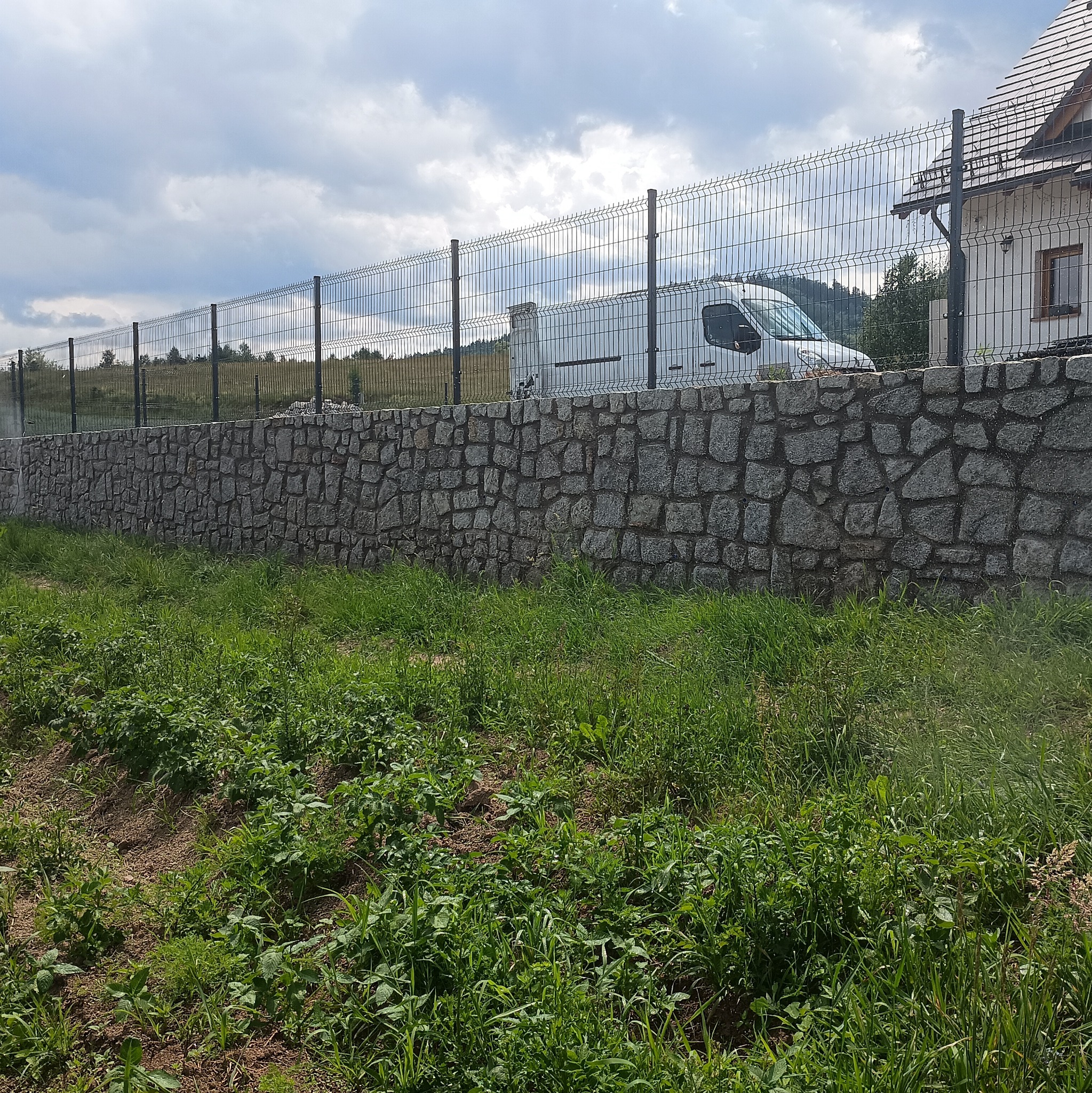 Granitowa podmurówka z metalową siatką ogrodzeniową, za nią częściowo widoczny biały budynek i zaparkowany biały samochód dostawczy, na pierwszym planie zarośnięty teren.