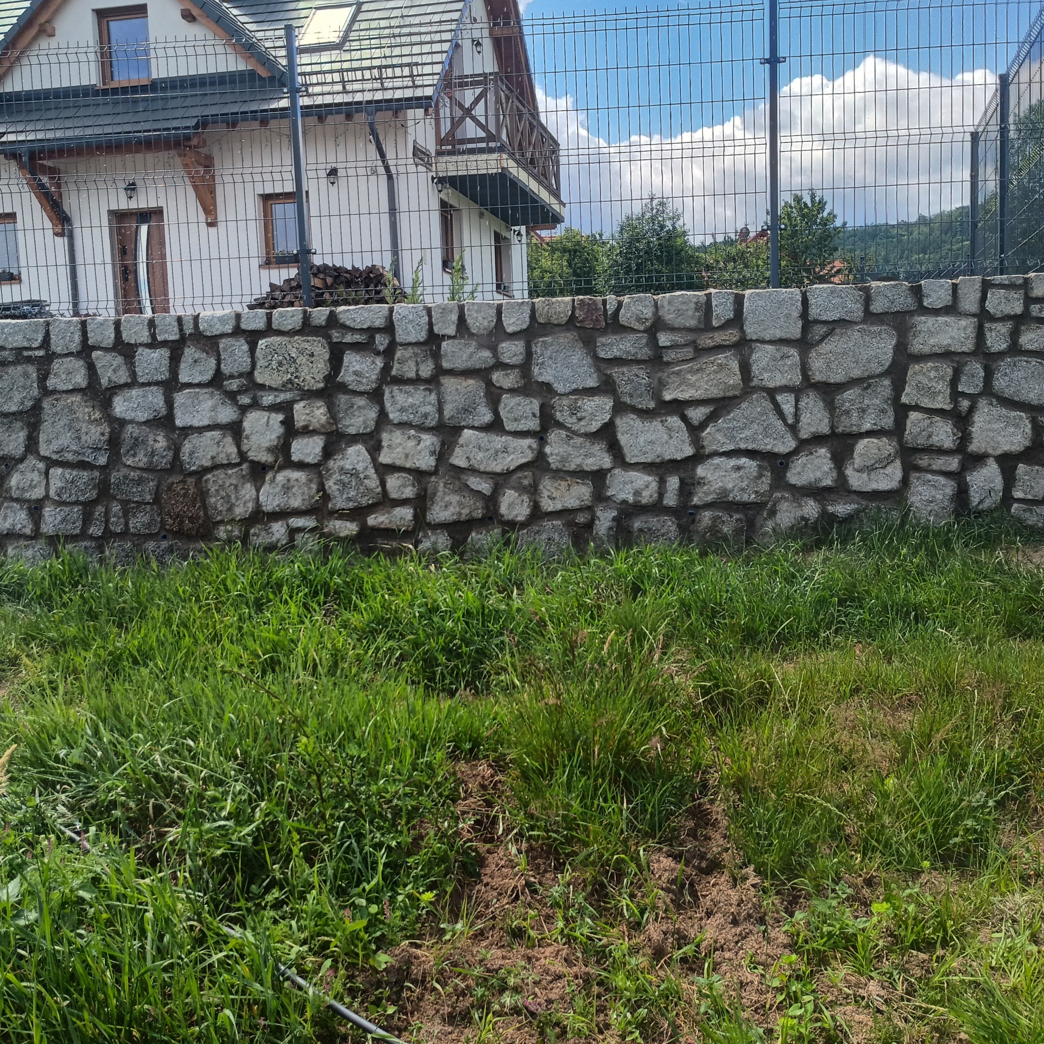 Murek z nieregularnych kamieni granitowych przed domem z balkonem, za ogrodzeniem z siatki.